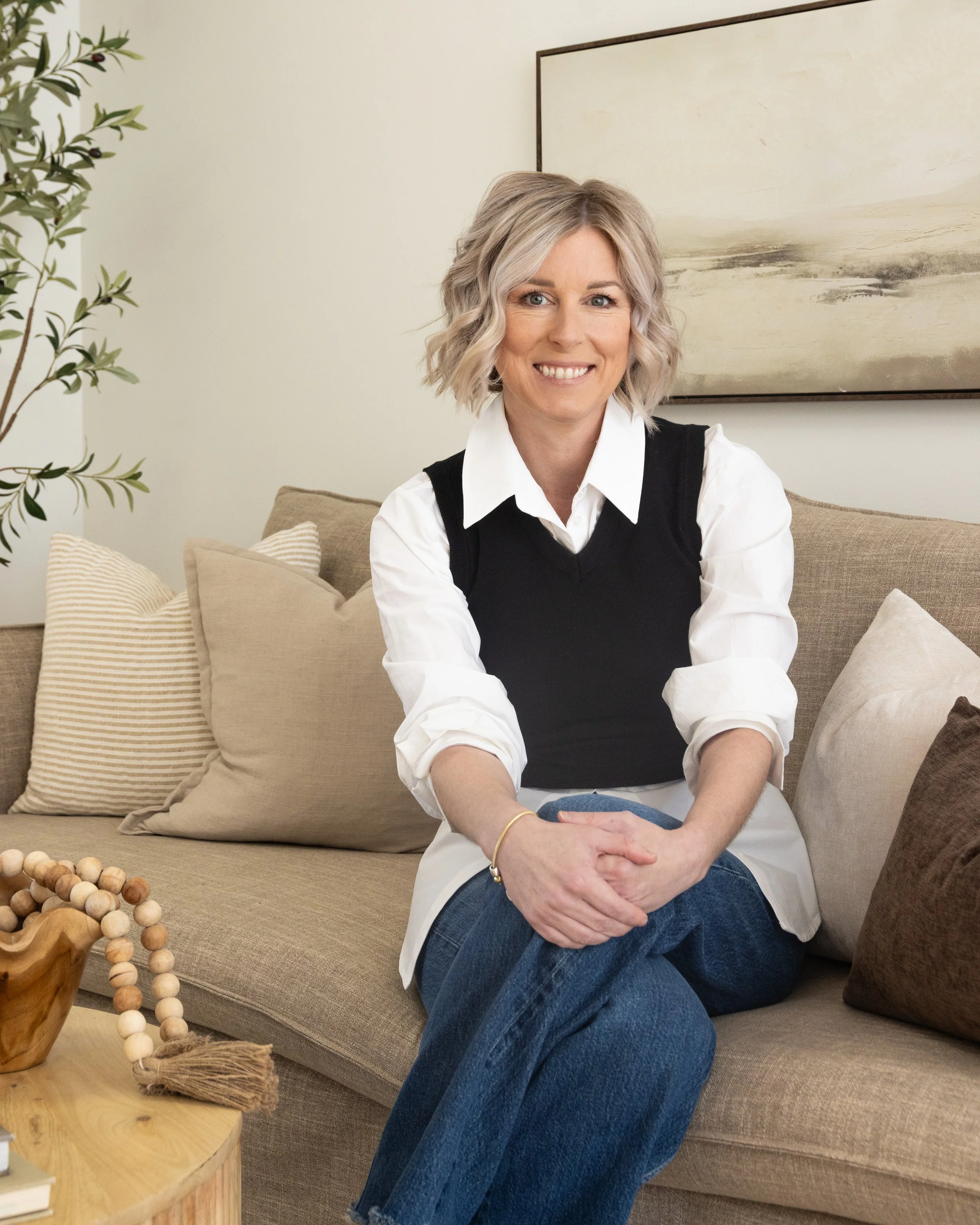 A woman with blonde, wavy hair sitting on a beige sofa in a living room, smiling at the camera. She is wearing a white shirt with rolled-up sleeves, a black vest, and blue jeans. There are multiple beige and brown pillows behind her and a framed abstract painting on the wall. A small round wooden table with decorative beads and a tassel design is in front of her. A potted plant is visible on the left side of the image.