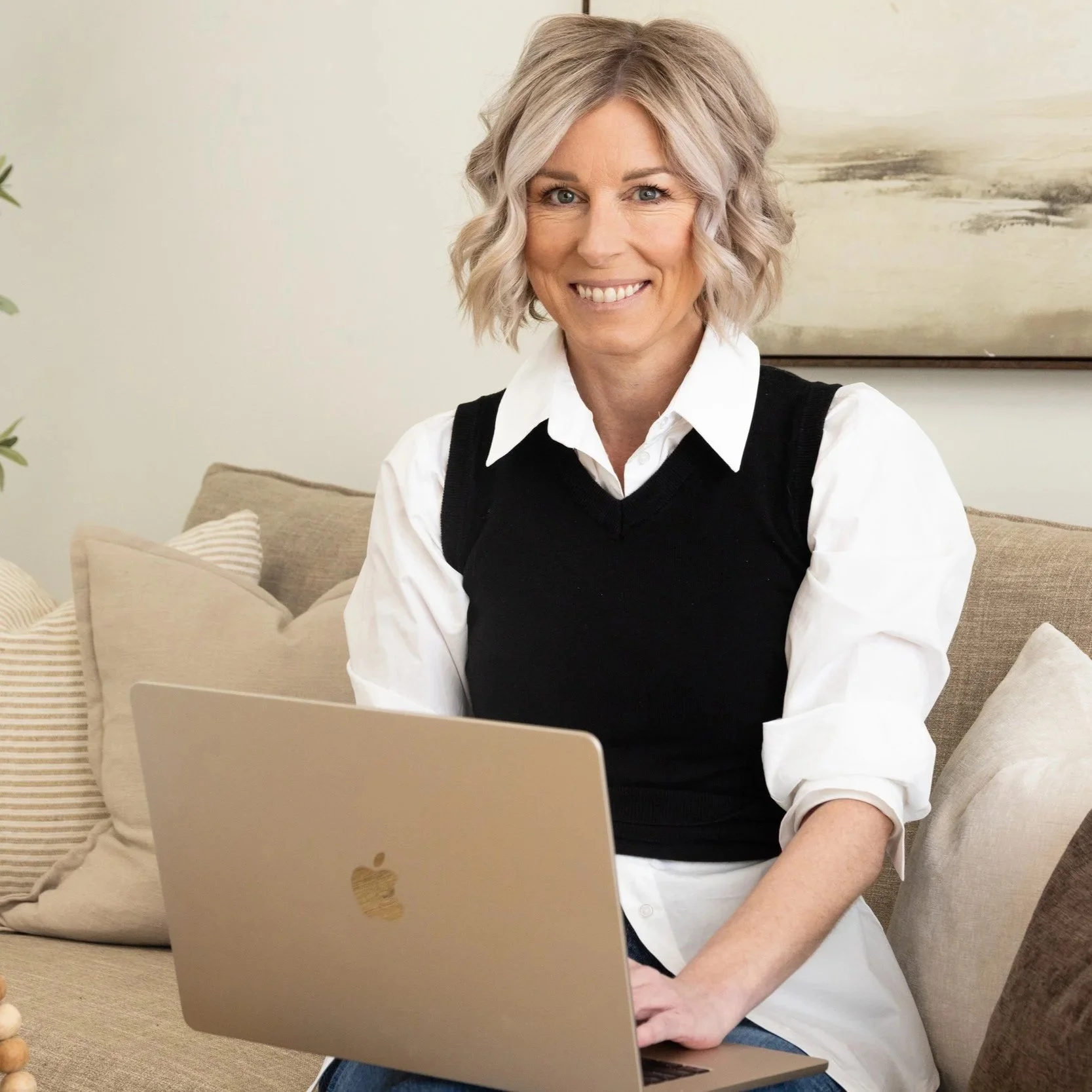 A woman with blonde, wavy hair sitting on a beige sofa, smiling at the camera with a silver Apple laptop on her lap in a home setting.
