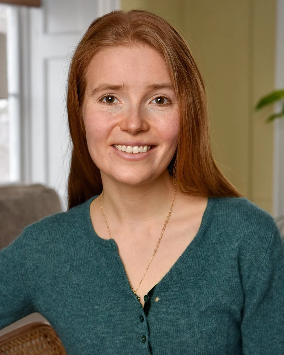 Close-up of a smiling woman with red hair, wearing a teal cardigan, sitting indoors near a window with a blurred background.