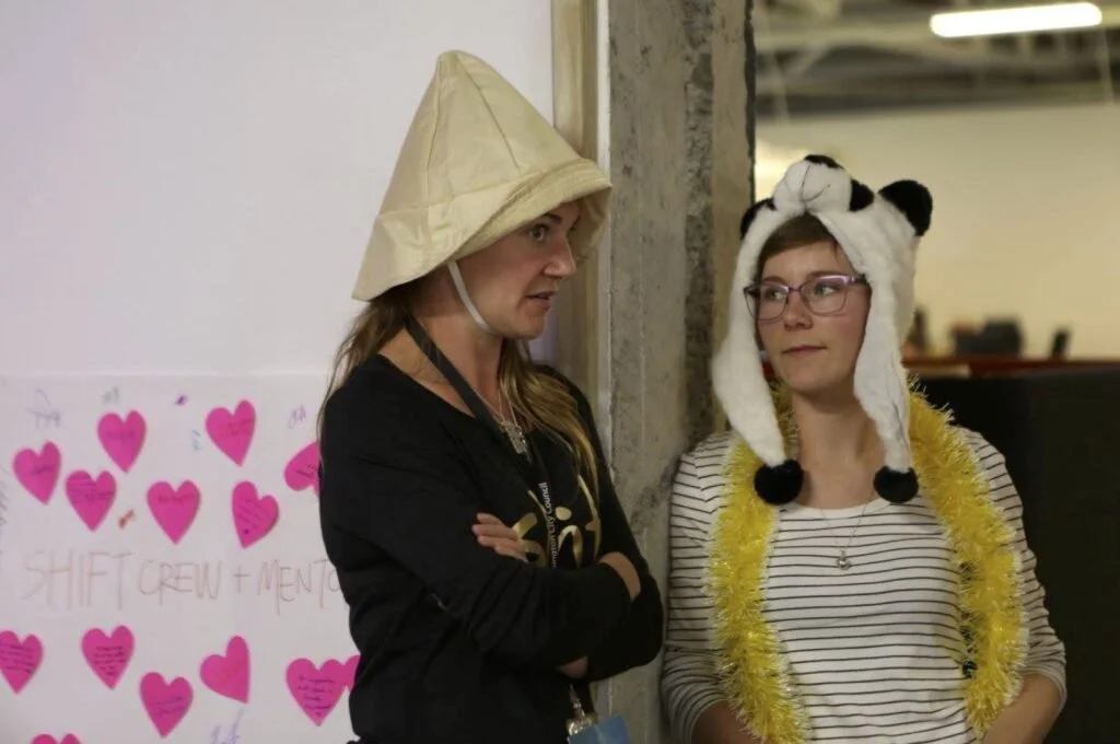 Two women standing indoors, one wearing a chimney sweep hat and the other wearing a panda hat and gold tinsel, engaged in conversation.