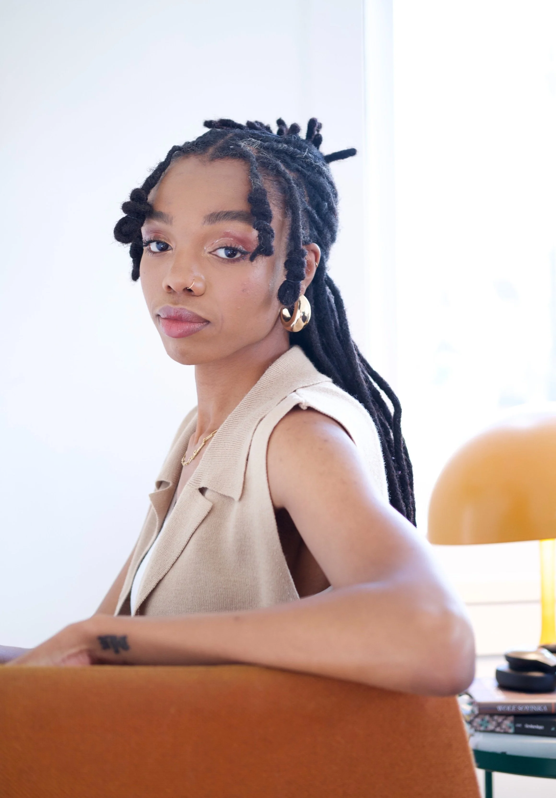 Soma Okoye is wearing a beige sleeveless top, gold hoop earrings, a necklace, and a nose ring, sitting at a desk in a bright room.