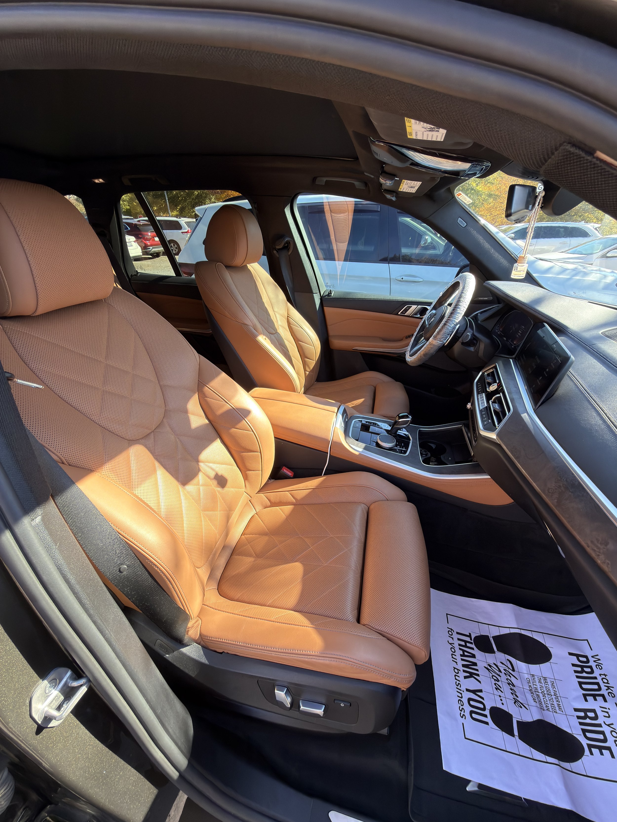 The interior of a modern car with tan leather seats, a black dashboard, and various controls. There is a paper floor mat with a thank you message for business.