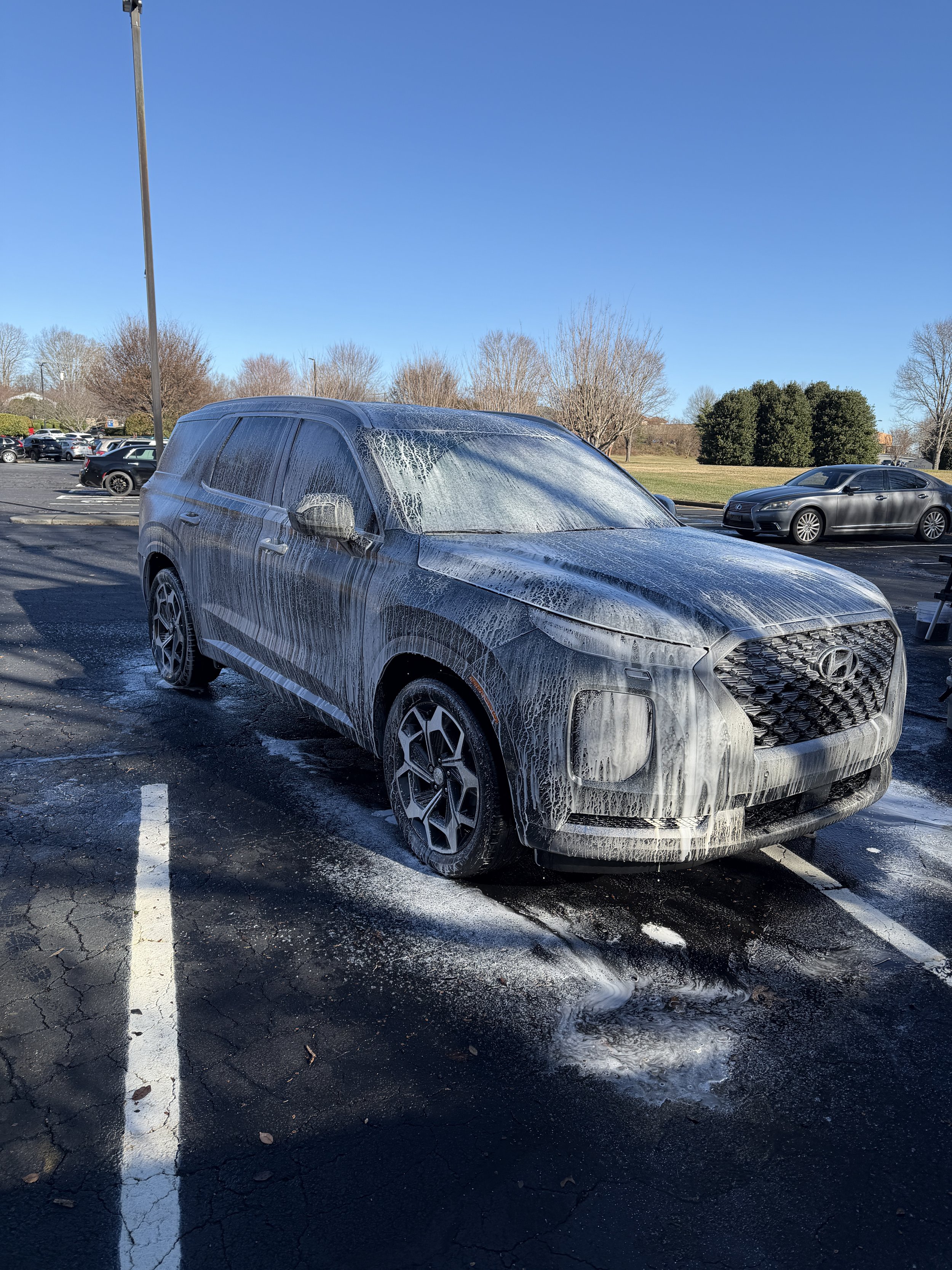 A black Hyundai SUV covered in white soap lather during a carwash in an outdoor parking lot under a clear blue sky.