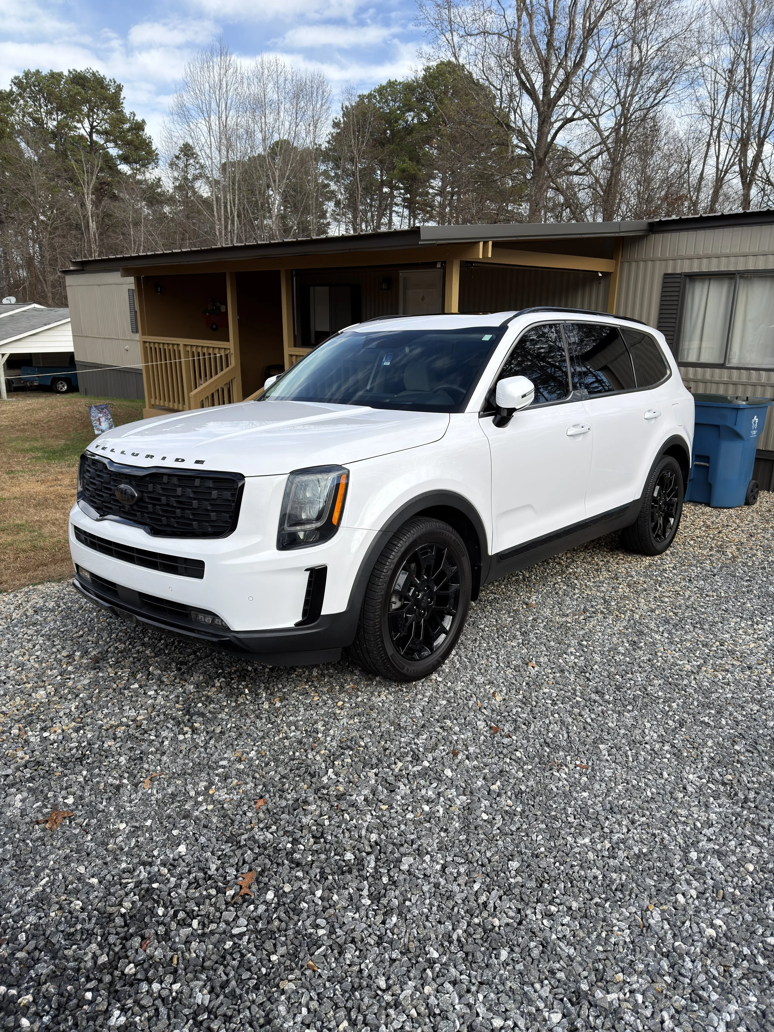 A white Kia Telluride SUV with black wheels parked on a gravel driveway in front of a house with a porch, surrounded by trees.