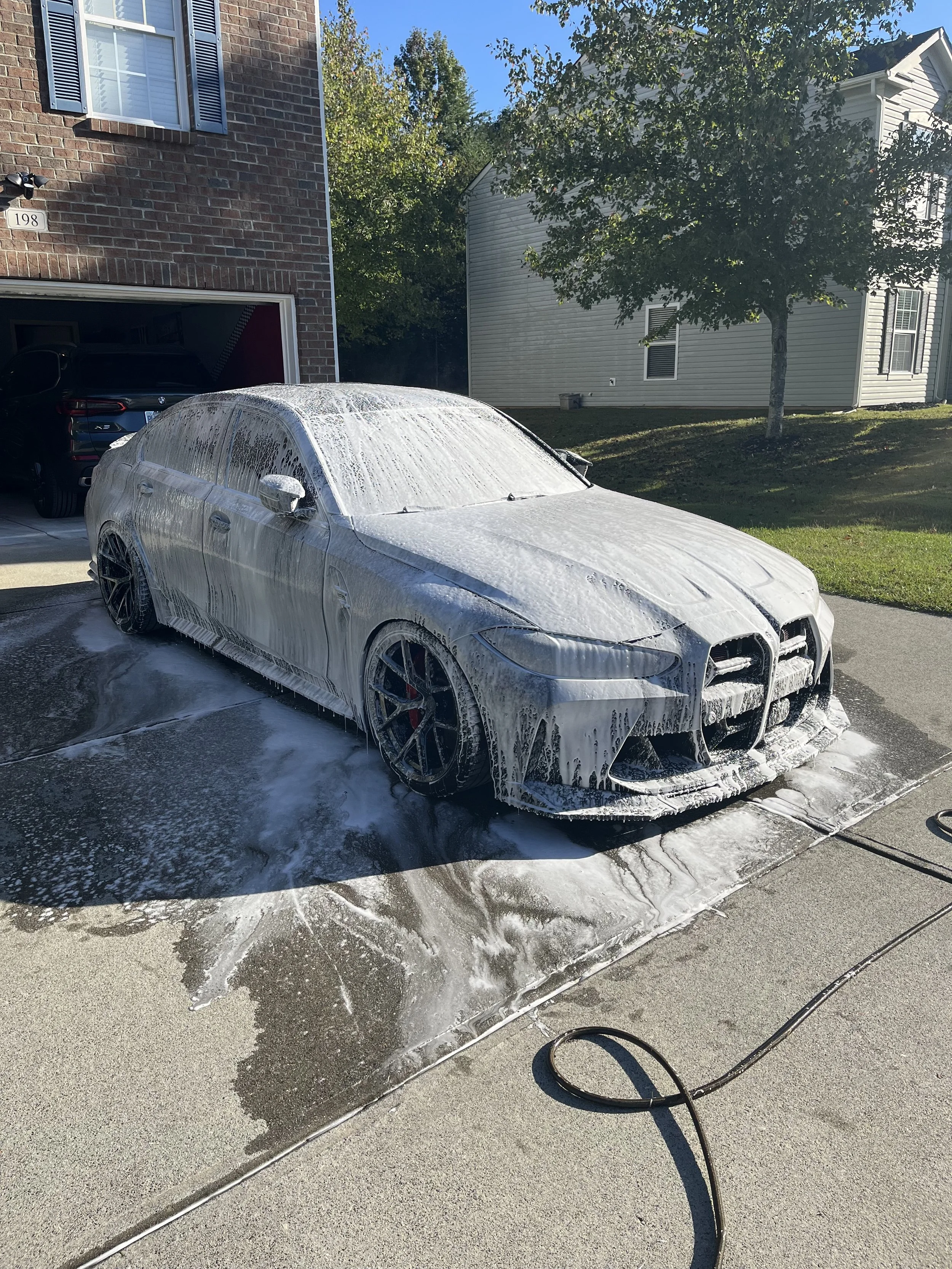 A silver sports car is being washed with foam on its surface in a driveway, with soap suds dripping onto the ground. In the background, a brick house and a white house are visible, along with a tree and a clear blue sky.
