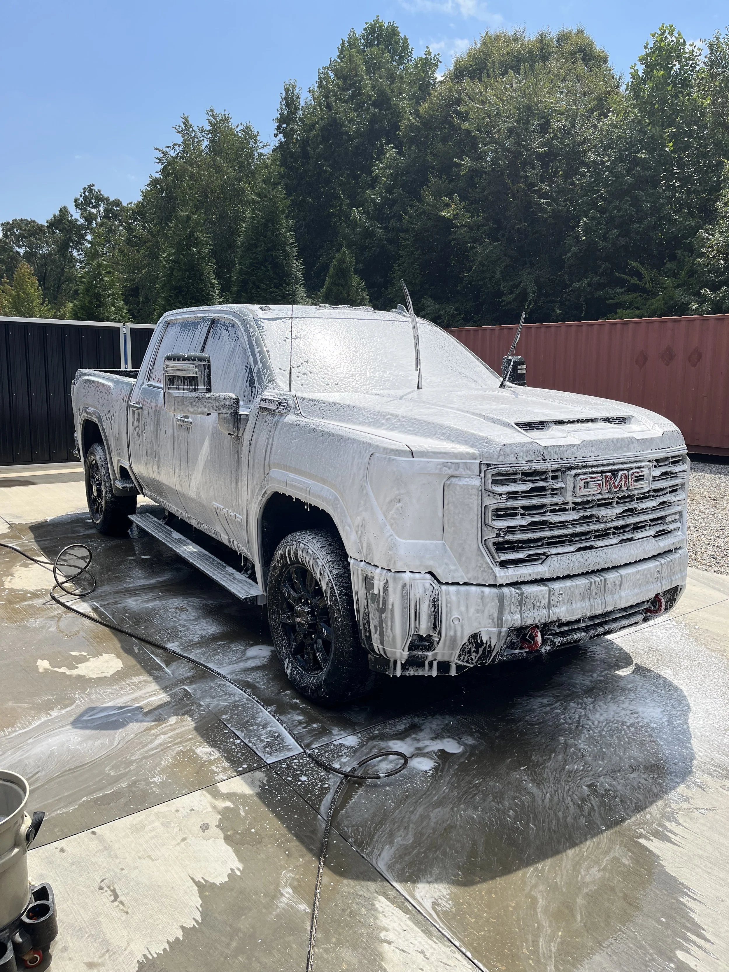 White GMC truck covered in soap suds during a wash, on a driveway with a garden hose nearby, with trees and shipping containers in the background.