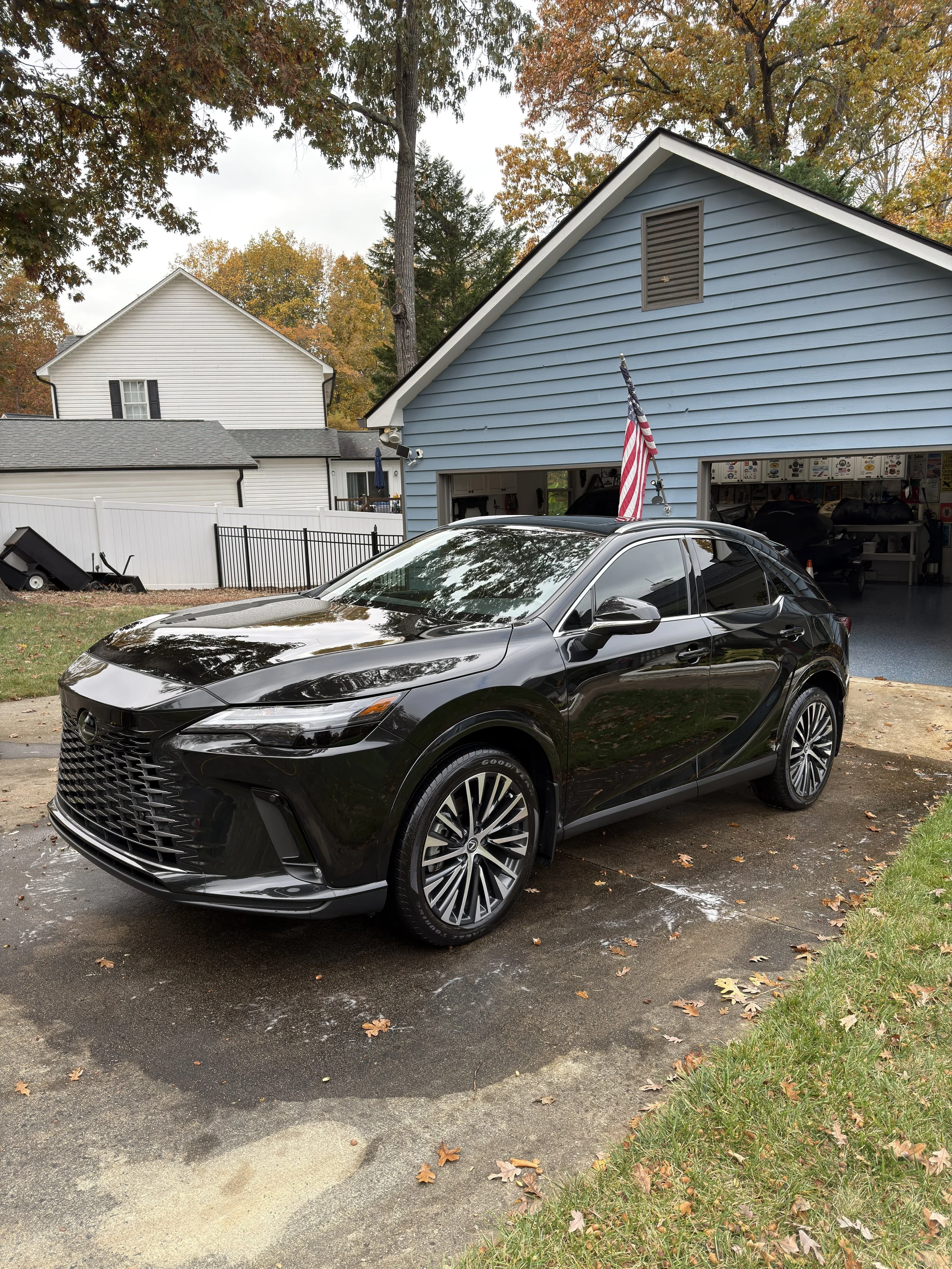 Black SUV parked in front of a blue garage with an American flag attached to it in a suburban driveway, with fall foliage in the background.