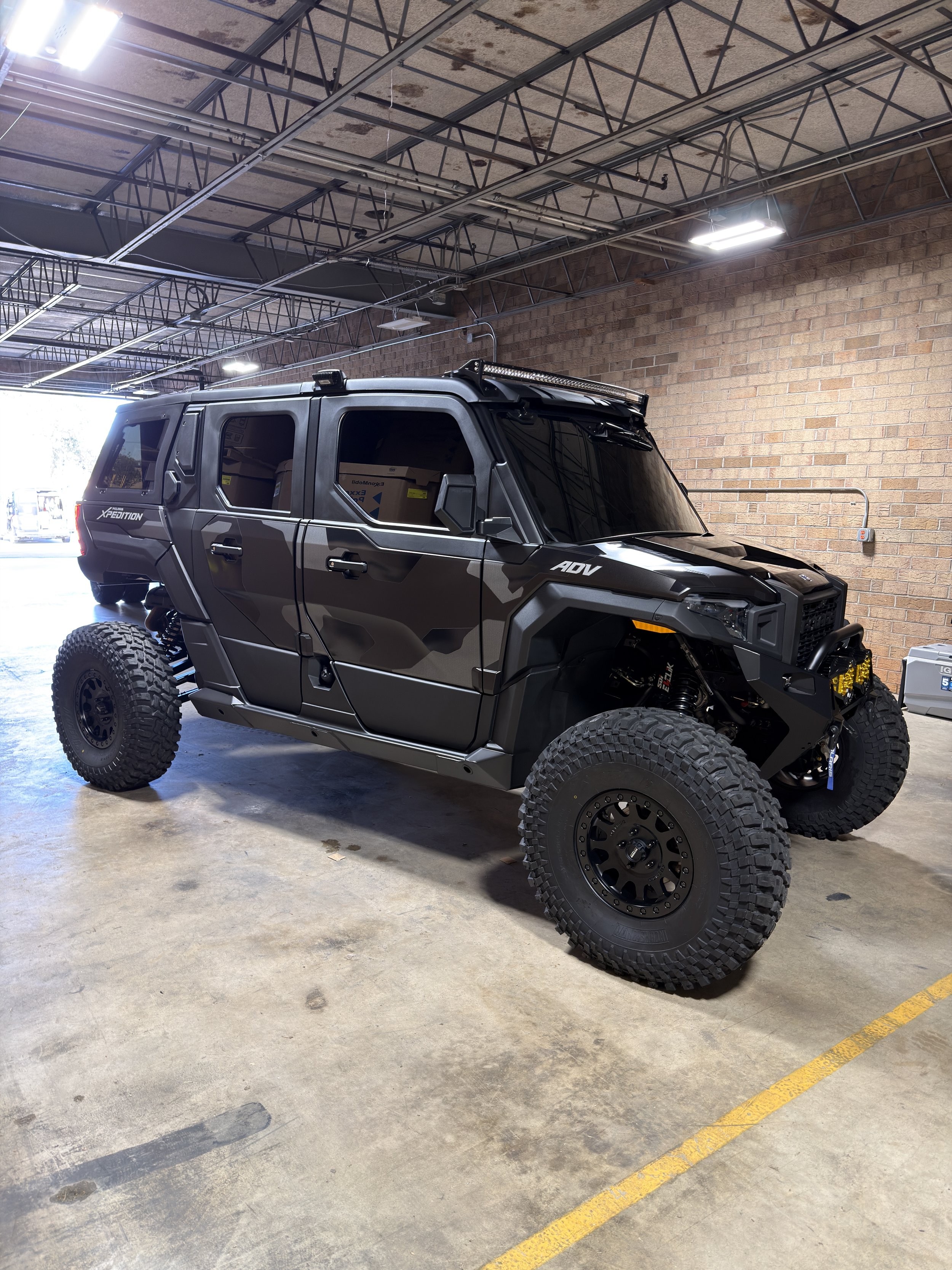 Black off-road utility vehicle with large tires parked in an industrial-style garage with brick walls and exposed ceiling beams.
