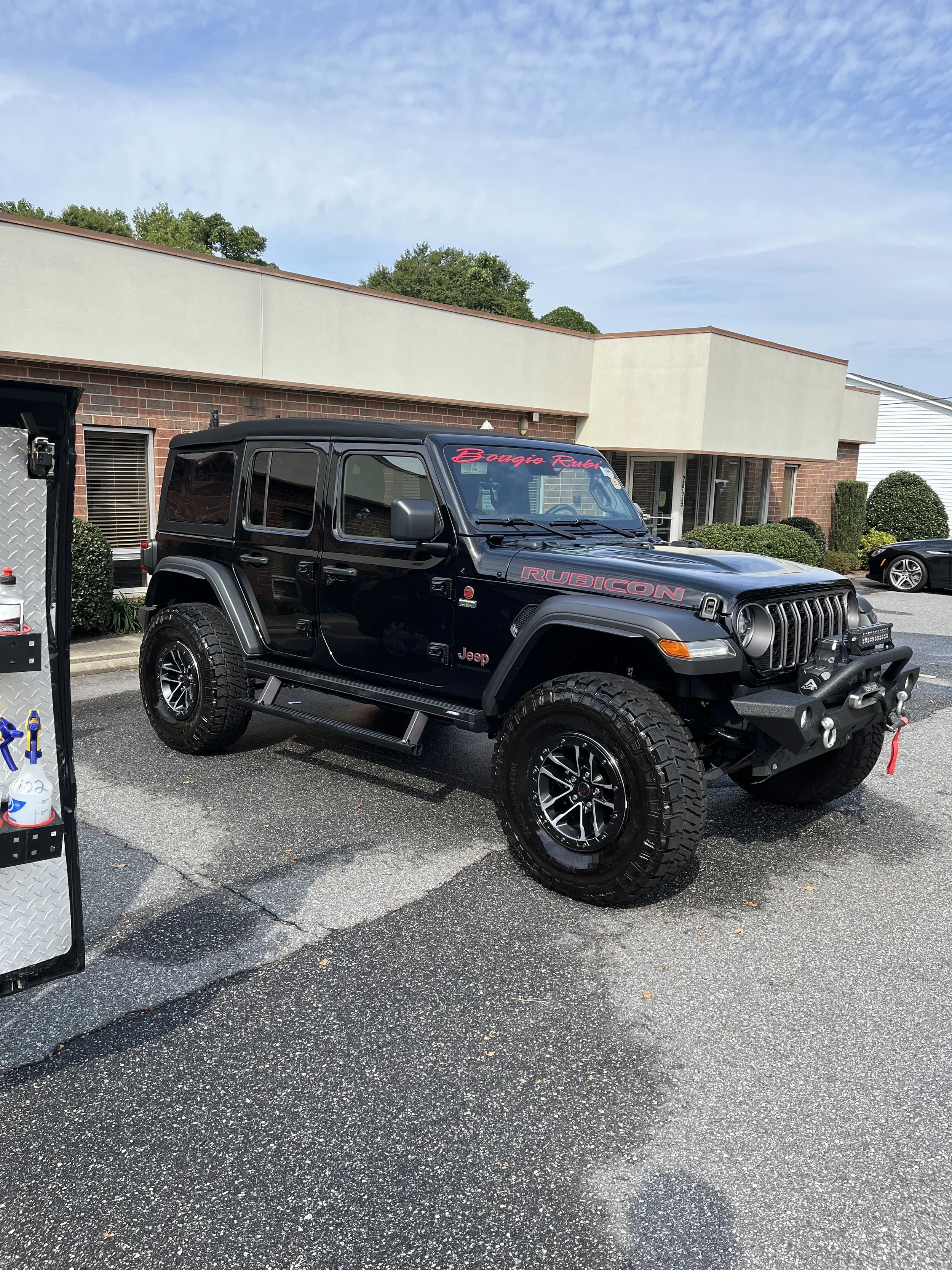 Black Jeep Rubicon parked in a parking lot next to a building with bushes and another vehicle in the background.