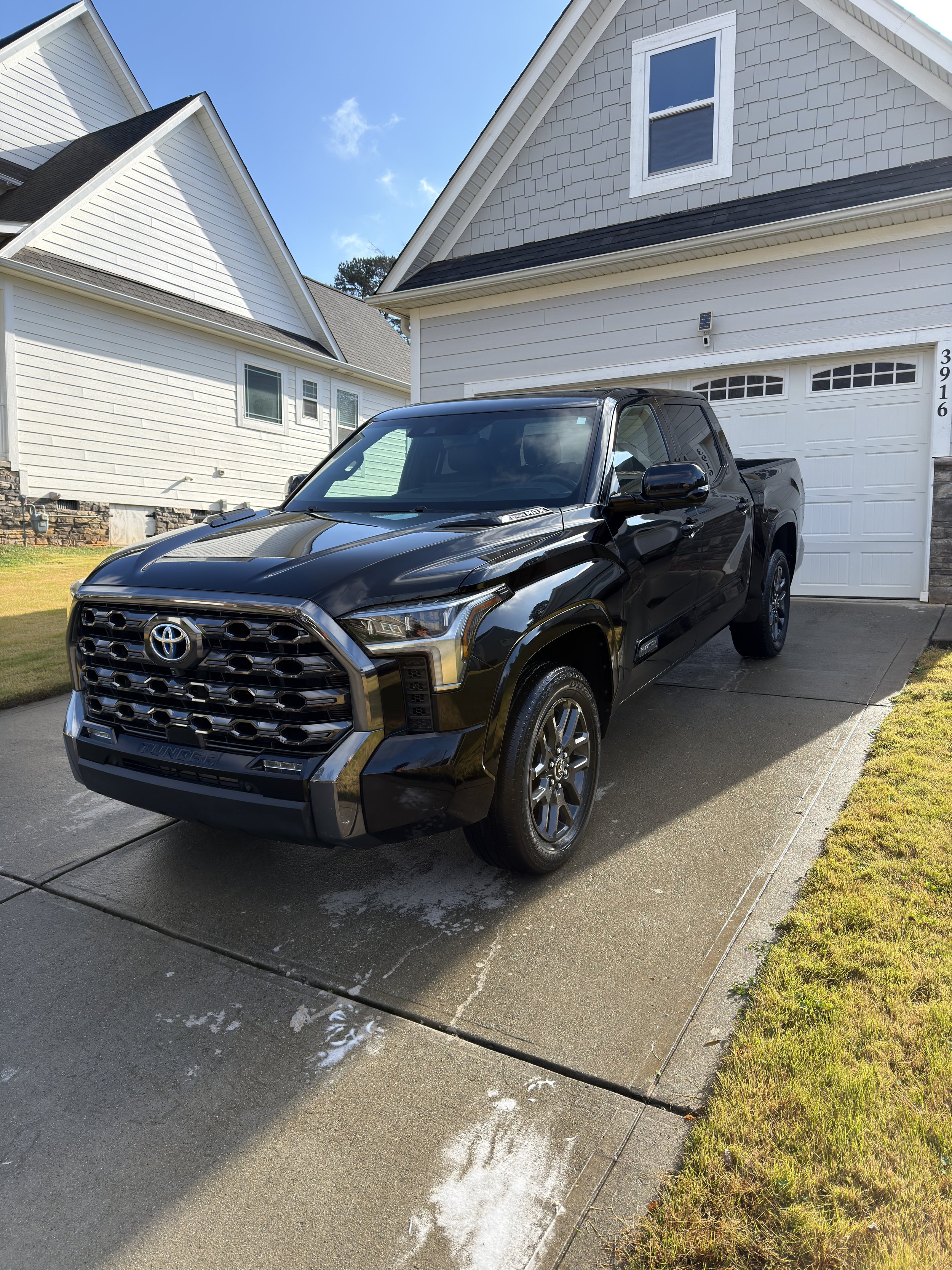 A black Toyota Tundra pickup truck parked on a concrete driveway in front of a white house with garage, under a clear blue sky.
