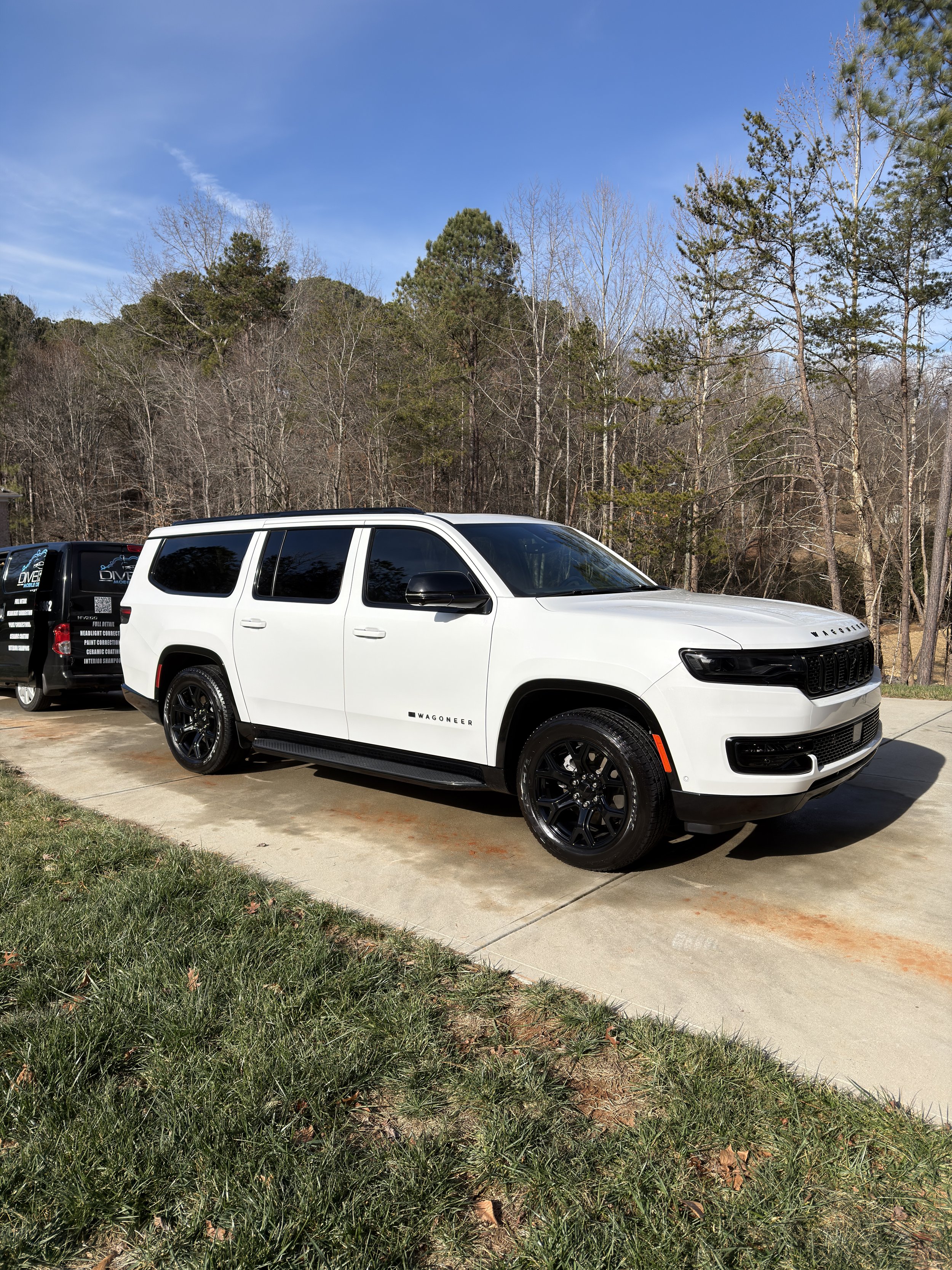 A white Jeep Wagoneer SUV parked on a concrete driveway with trees and a blue sky in the background.