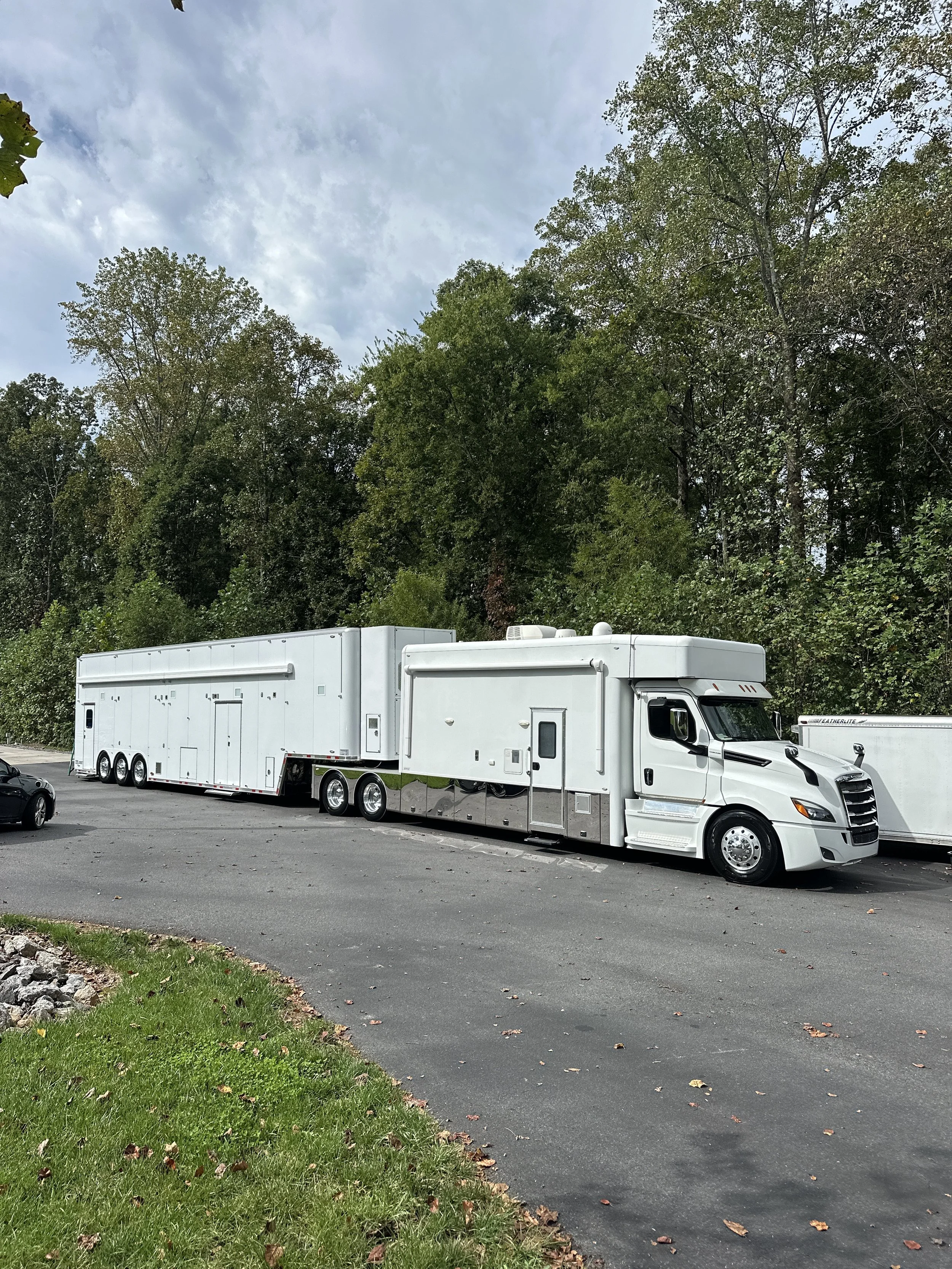 A large white truck with an attached trailer parked on a paved lot near a wooded area.