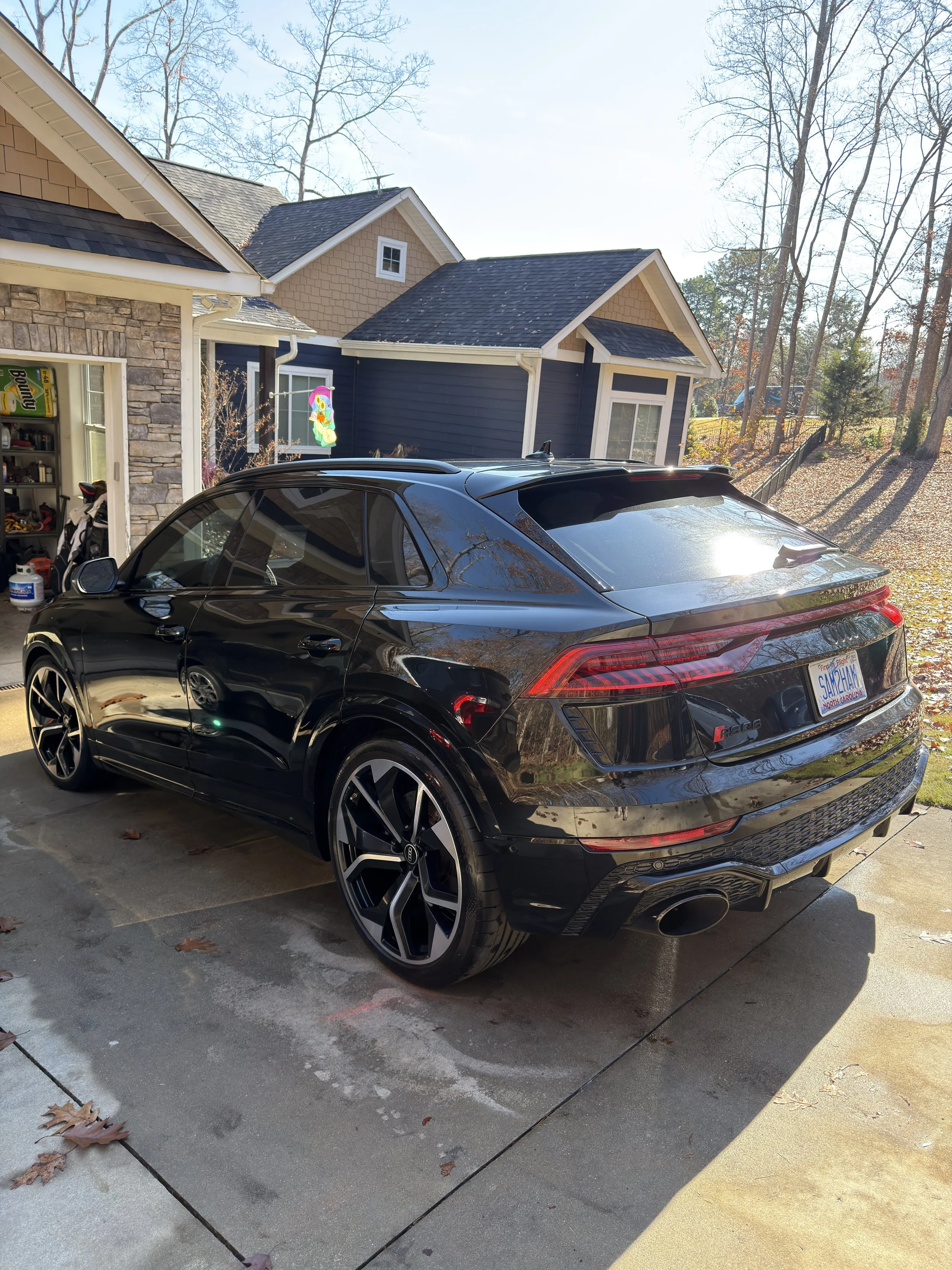 Black Audi SUV parked in a driveway outside a residential house with a blue and beige exterior and a garage with garden tools inside.