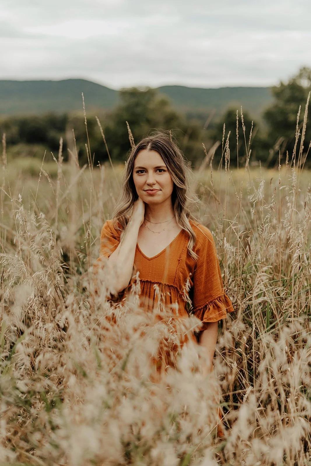 A young woman with wavy brown hair in an orange dress standing in a field of tall grass with mountains and cloudy sky in the background.