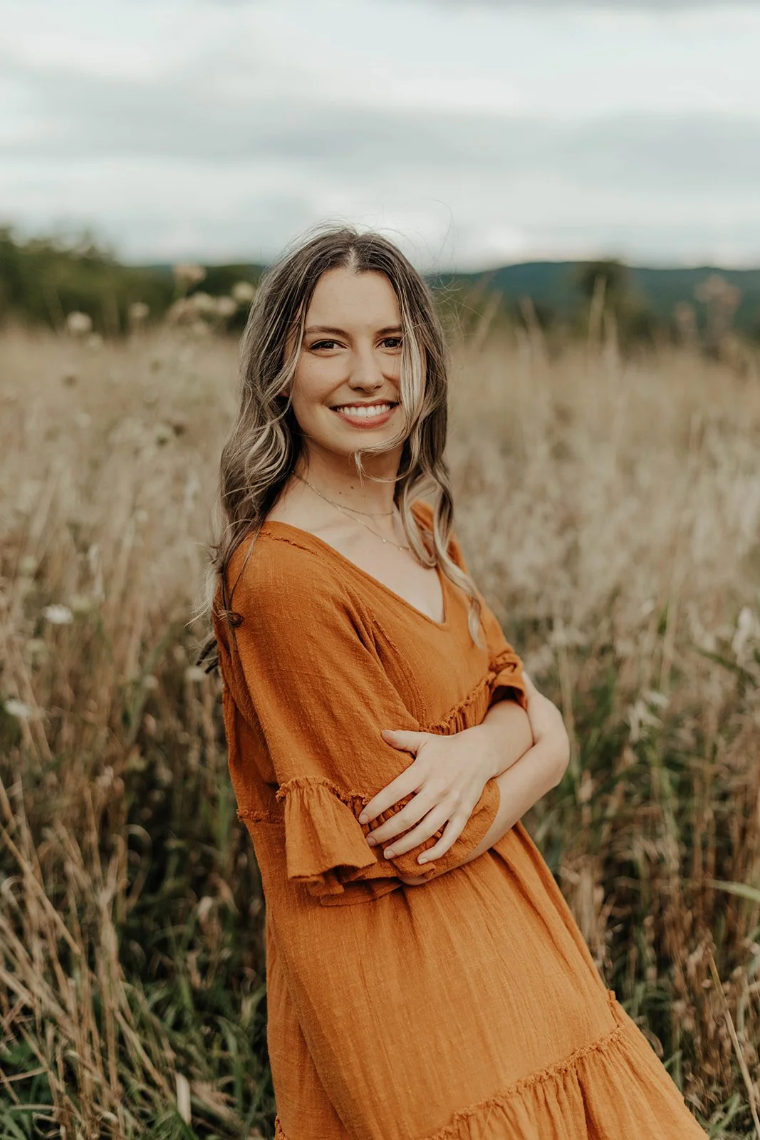 A woman with wavy hair smiling, wearing an orange dress, standing in a field with tall grass and mountains in the background.