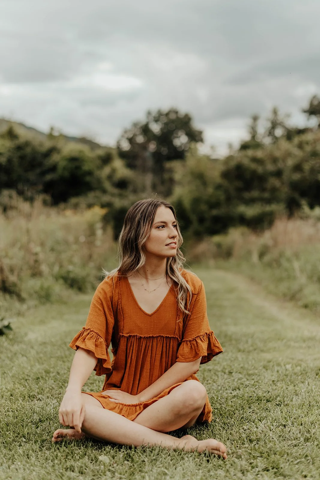 A woman with wavy hair sitting cross-legged on grass outdoors, wearing an orange dress with ruffled sleeves, surrounded by trees under a cloudy sky.