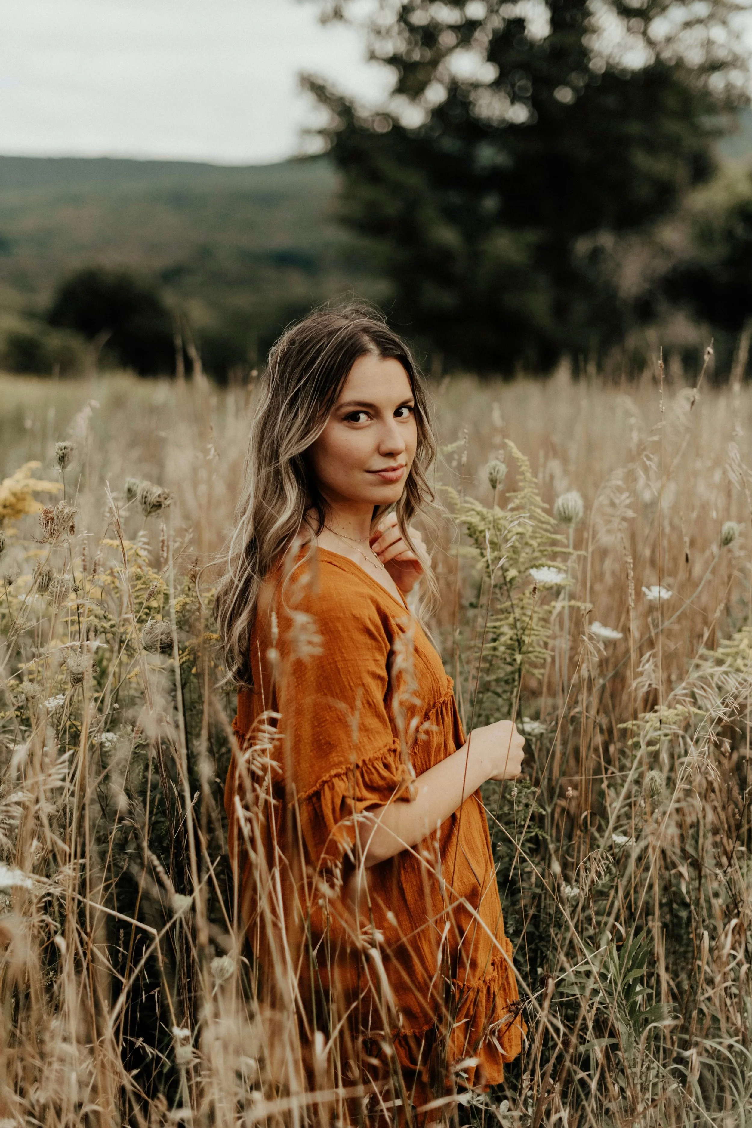 A woman with wavy brown hair in an orange dress standing in a field of tall grasses and wildflowers with trees and hills in the background.