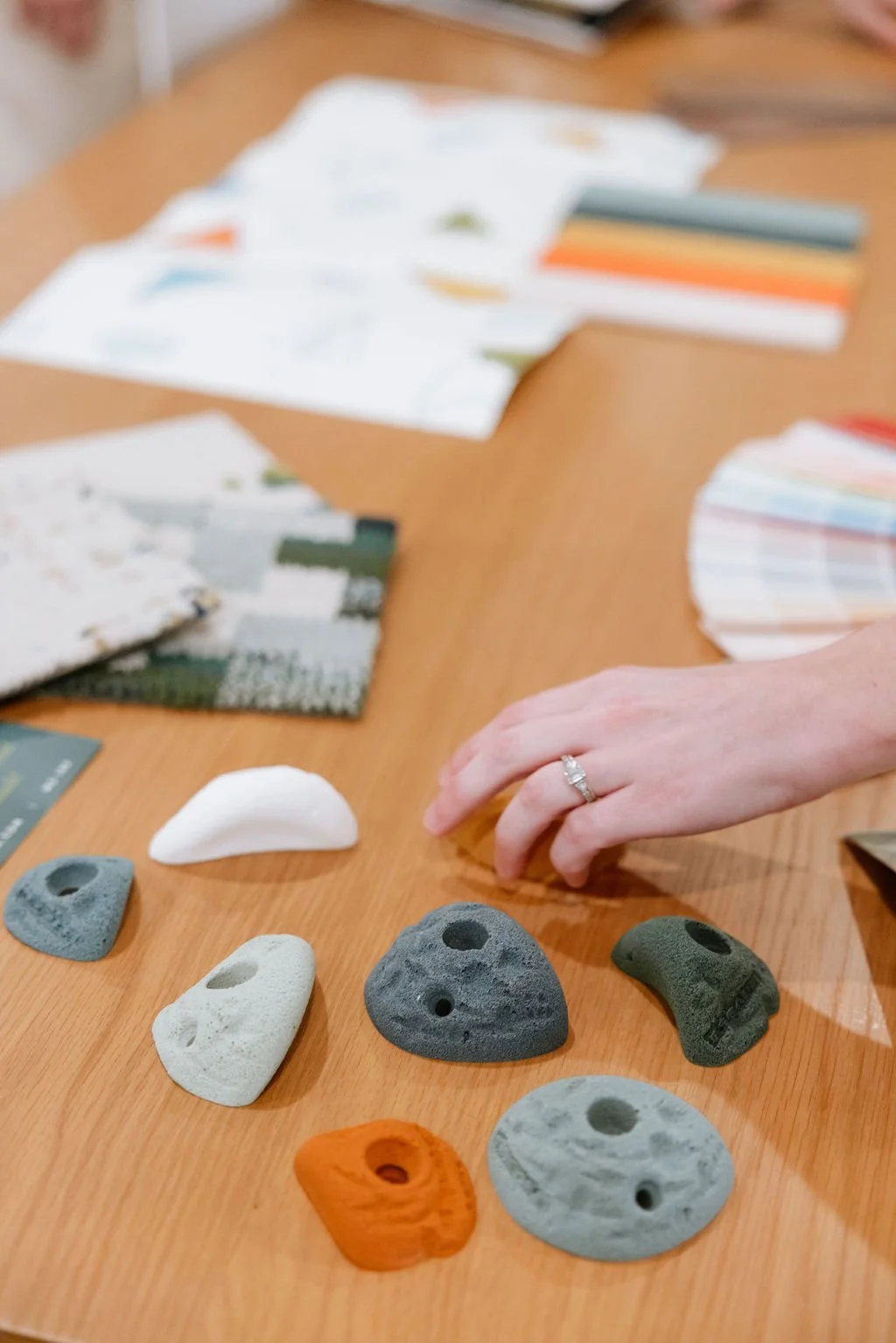 A hand with a ring reaches for a rock climbing hold on a wooden table, with several other climbing holds in various colors and shapes laid out nearby.