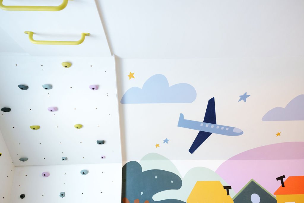 Indoor climbing wall with colorful holds and a painted sky mural featuring clouds and an airplane.
