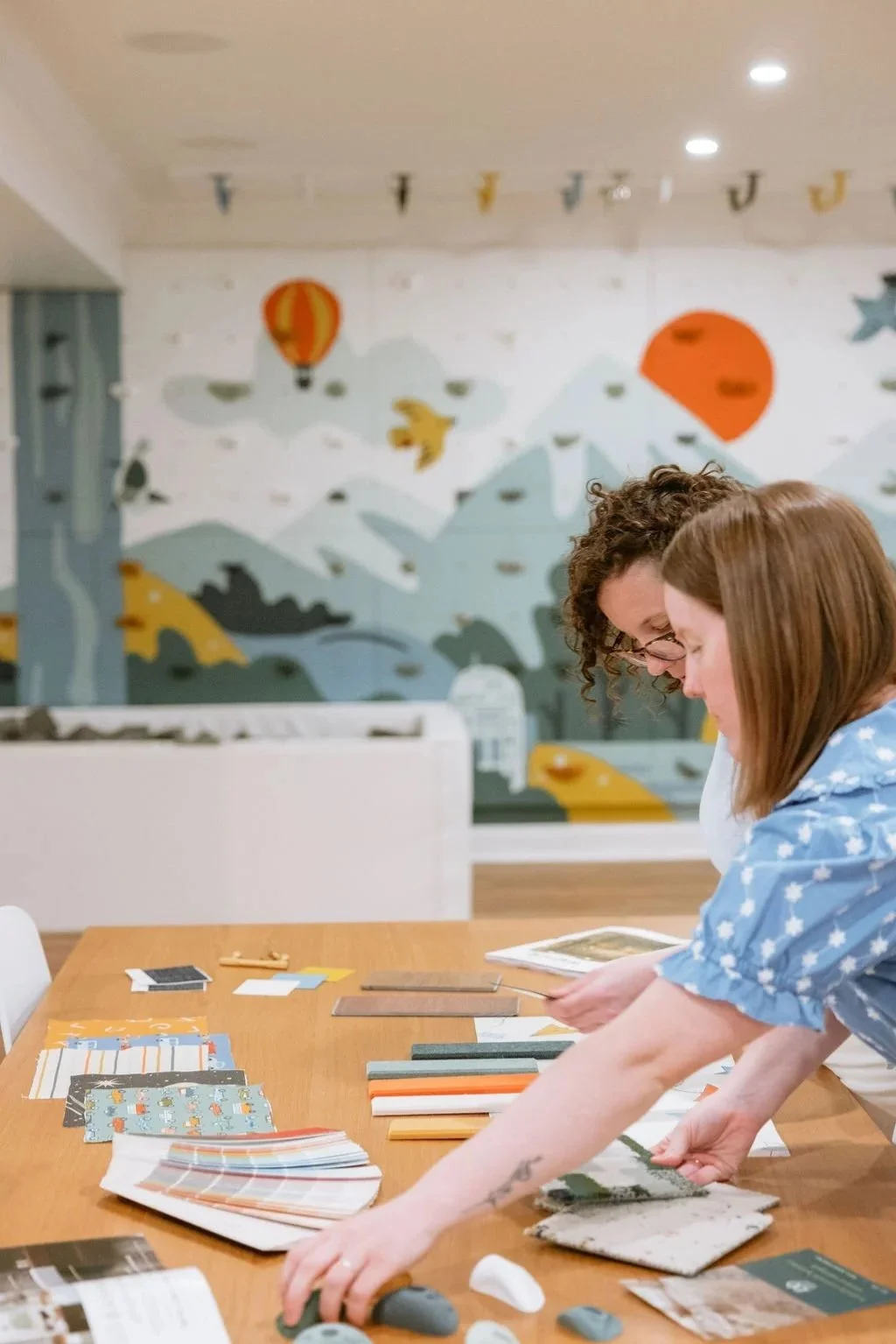 Two women looking at paint or wallpaper sample swatches on a table in an art studio decorated with a mountain scene mural and sky-themed bunting.