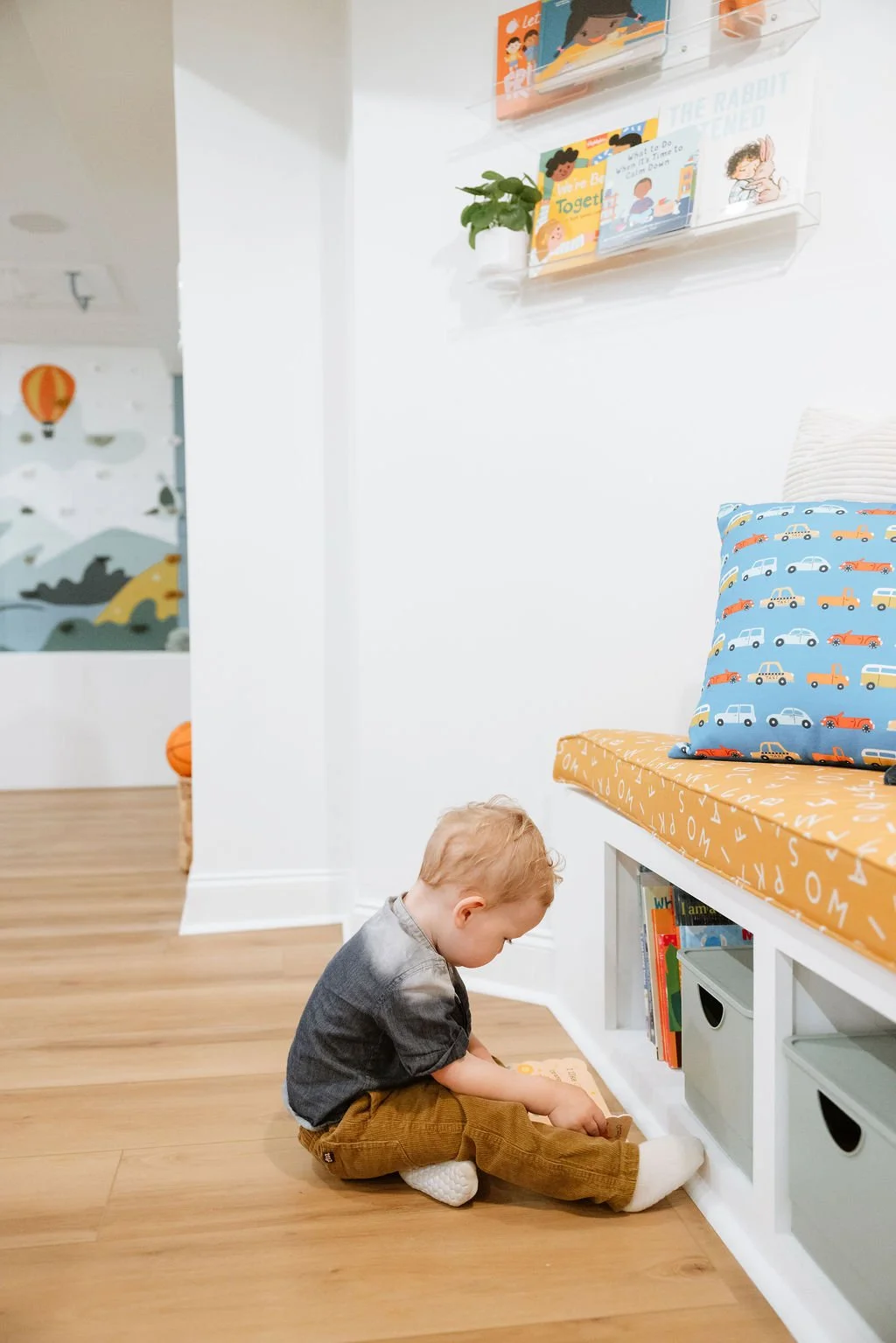 A young child with blonde hair sitting on a wooden floor, playing with a book by a white bookshelf with baskets. Behind them is a bench with a blue pillow decorated with cars. The wall above features children's books and a plant, with a mural of a hot air balloon and animals in the background.