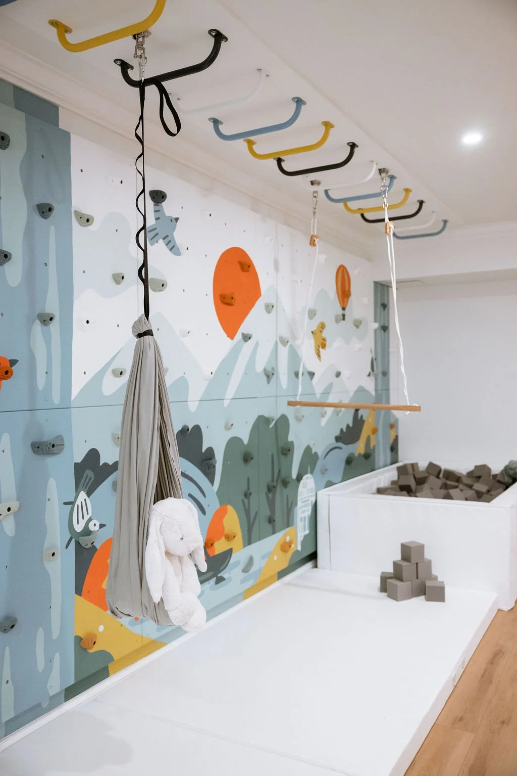Custom playroom with a rock wall, monkey bars, sensory swing, trapeze bar, foam pit and landing mats, There is a custom mural in the background.