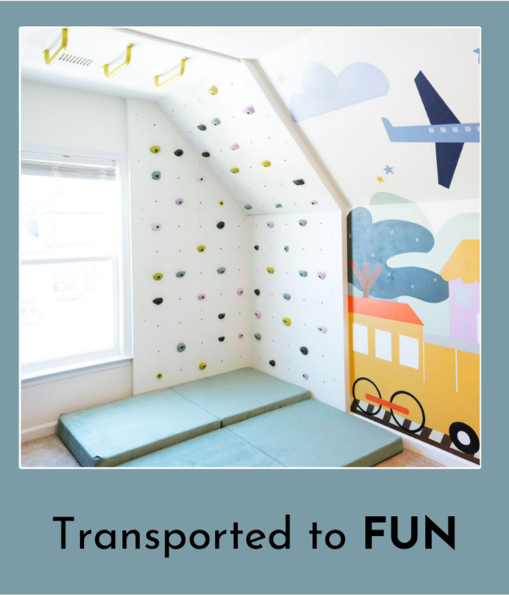 A small indoor climbing wall with colorful holds, padded mats on the floor, a nearby window, and a mural of a train, clouds, and an airplane on the wall.