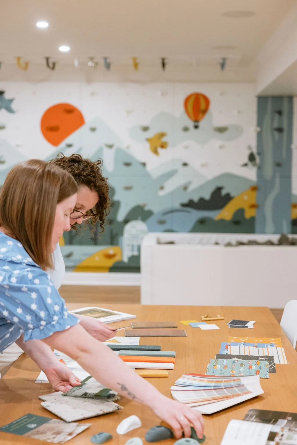 Playroom designers, Kayla and Kristin looking through fabric samples and design materials on a table in front of a customised rock wall and foam pit.