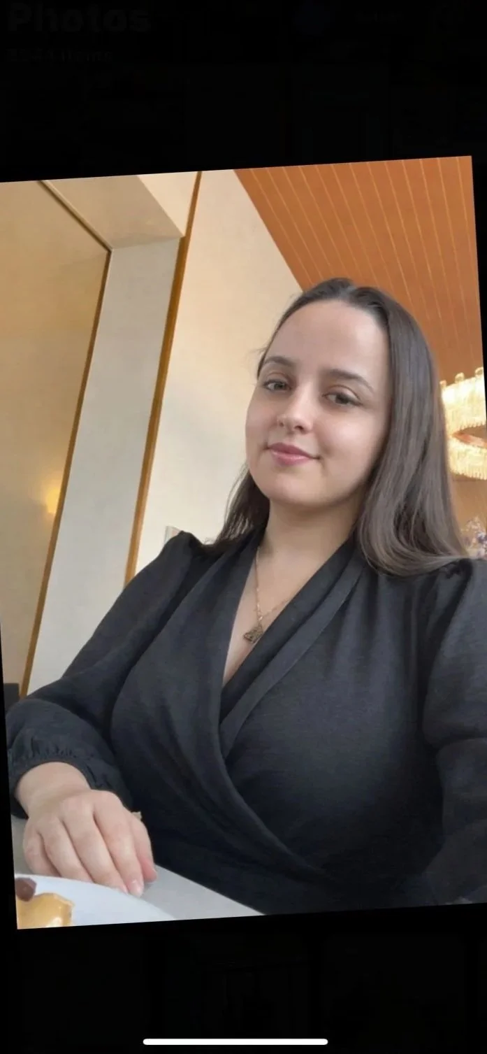 A young woman with long brown hair sitting at a table, smiling slightly, wearing a black wrap-style top, in a modern indoor setting with warm lighting and wooden ceiling panels.