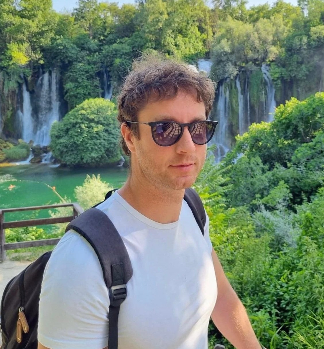 A young man with dark hair and sunglasses standing outdoors in front of green trees and waterfalls.