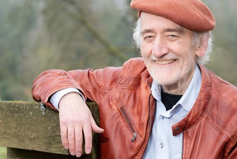 Älterer Mann mit roter Jacke und Mütze lehnt lächelnd an einer Holzbank im Freien