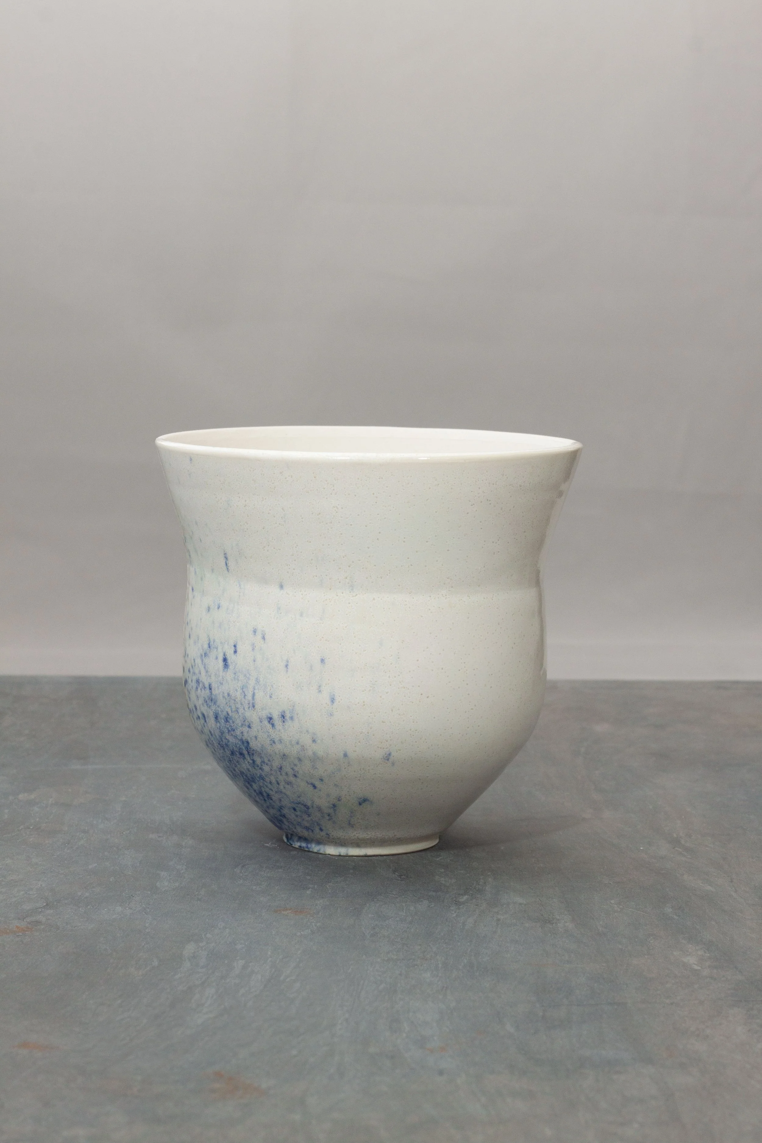 A white porcelain ceramic vase with a subtle speckled pattern, placed on a gray surface with a plain gray background.