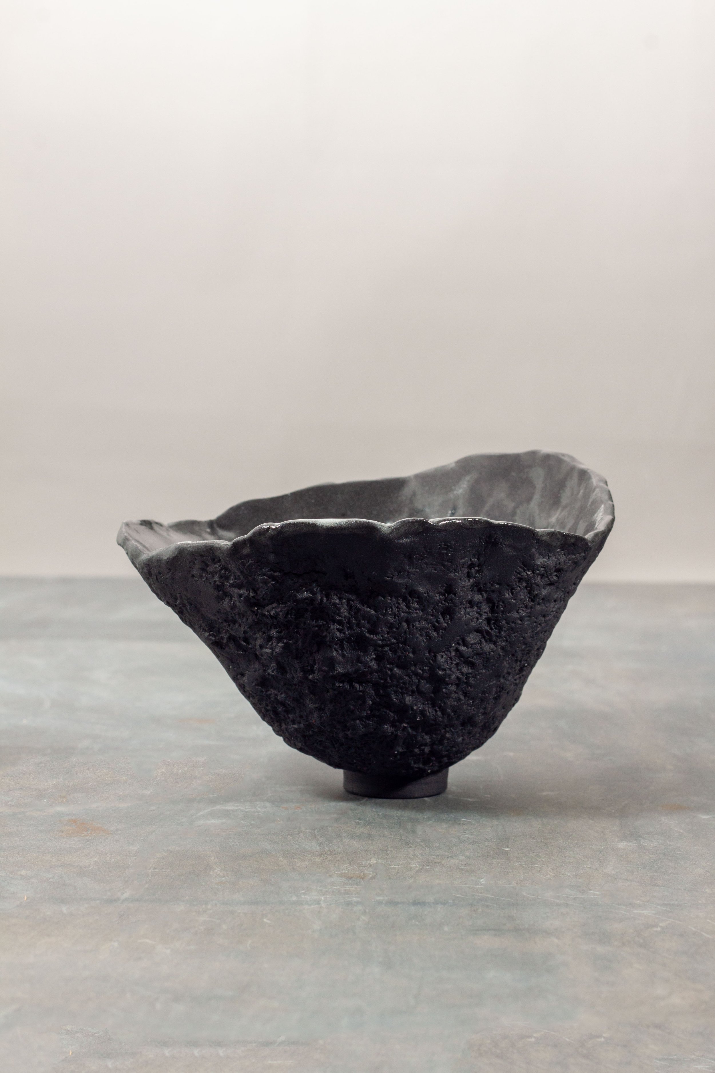A black porcelain ceramic bowl with a rough, textured exterior and a crinkled, wavy rim, placed on a grey surface against a plain light background.