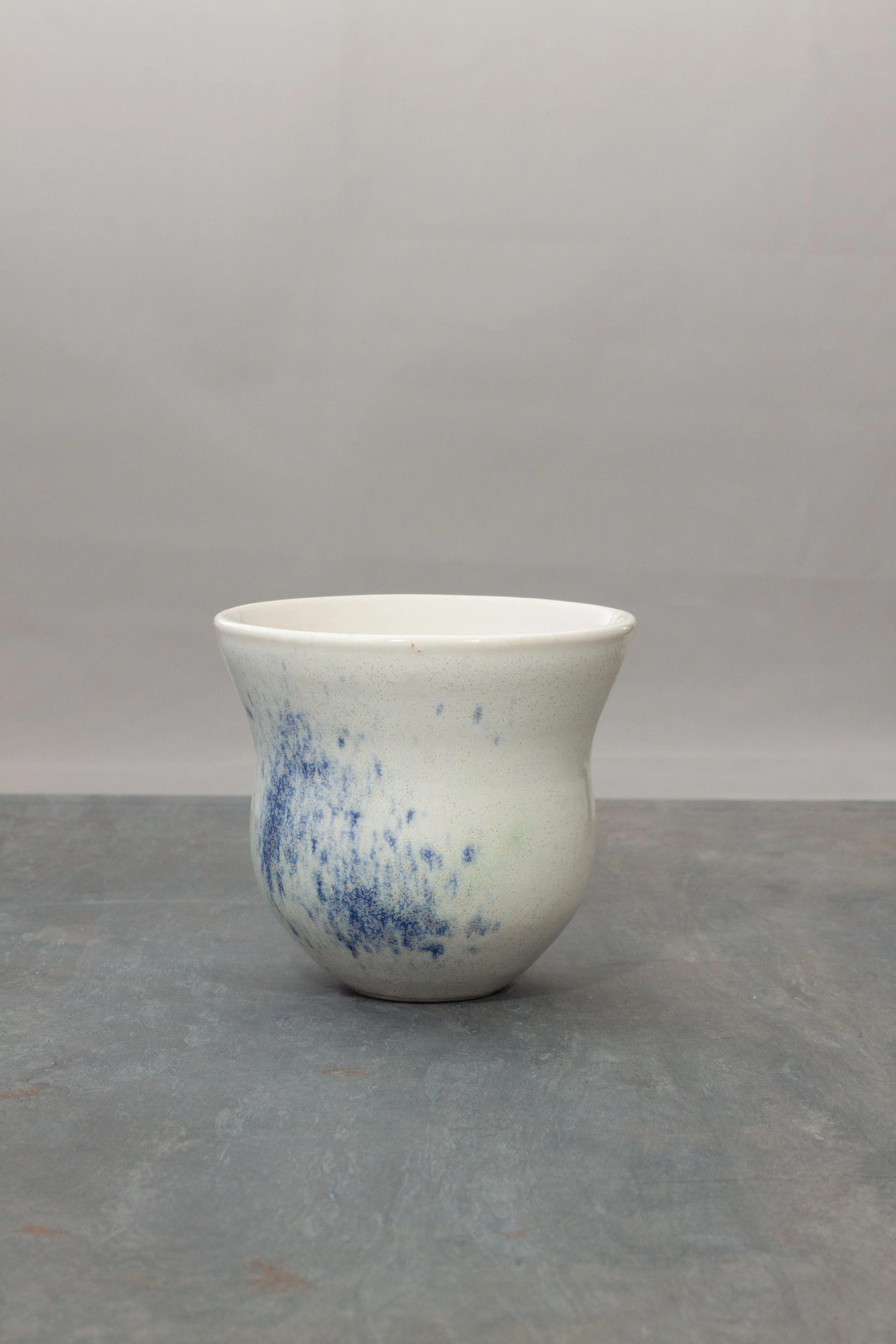 A white porcelain ceramic vase with blue speckles and a simple, curved shape, placed against a plain gray background.