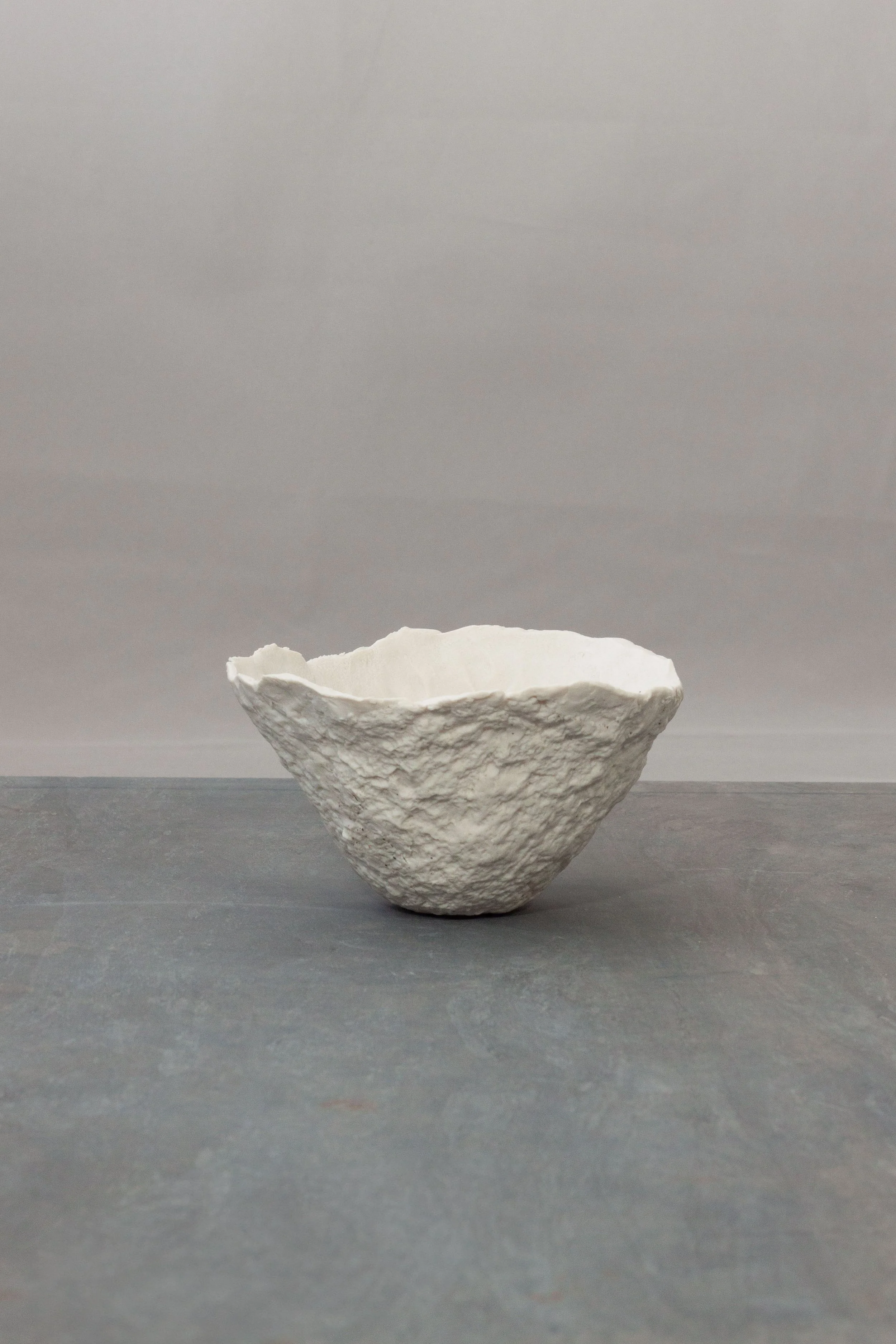 A small white porcelain ceramic bowl with rough, uneven edges and a textured exterior, placed on a gray surface against a gray background.