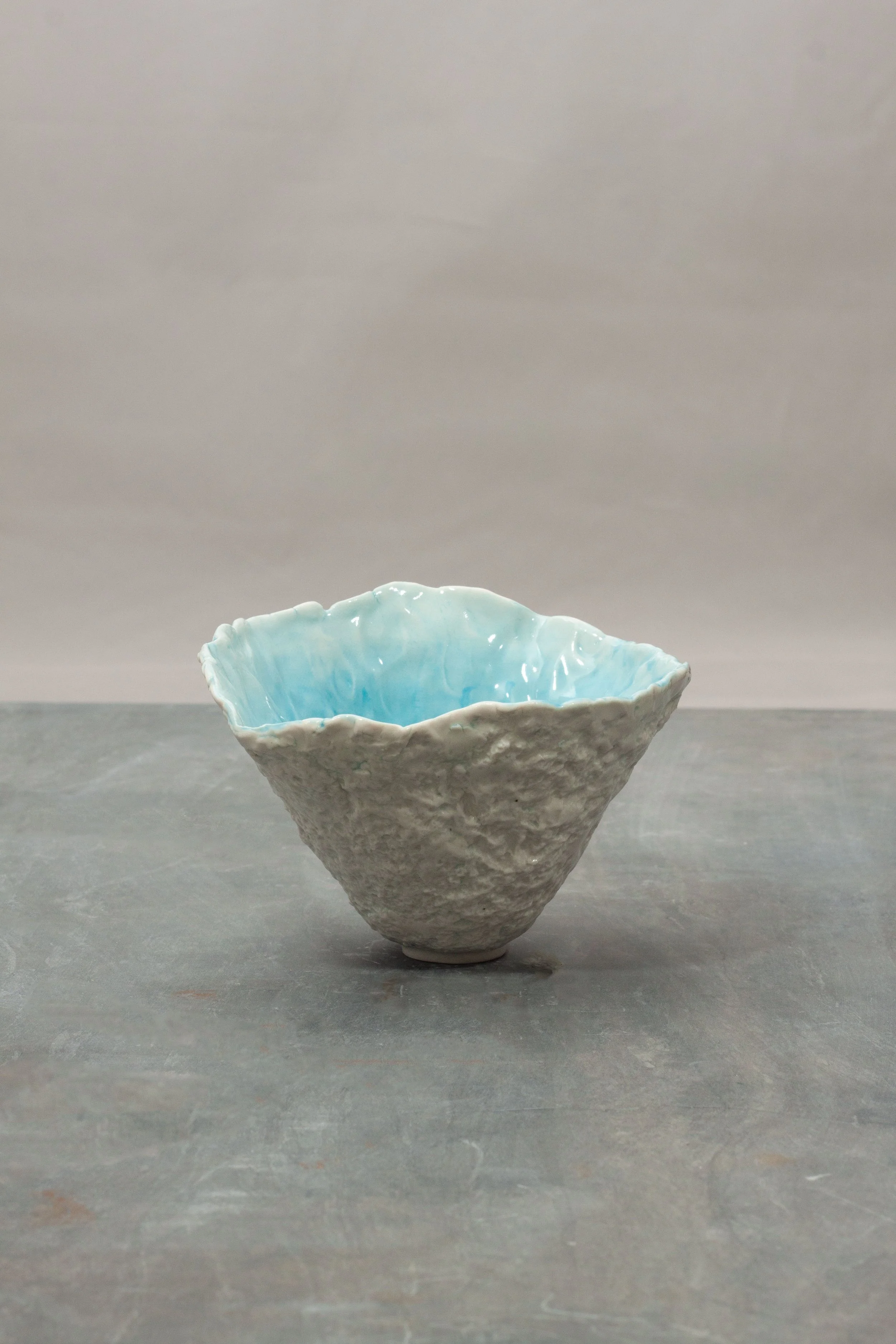 A small porcelain ceramic bowl with a rough, textured exterior and a glossy, light blue interior, placed on a gray surface against a plain, light gray background.