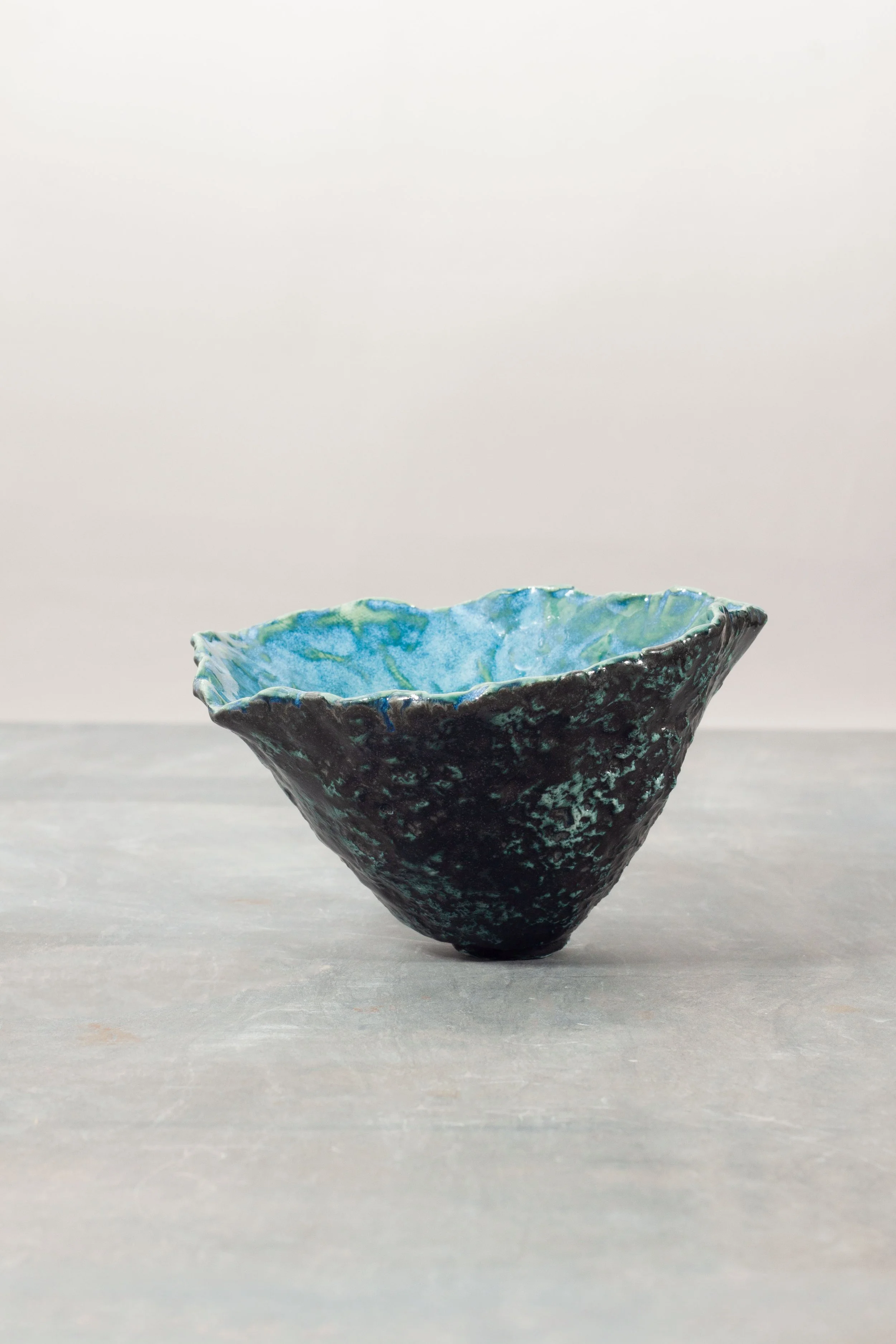 A dark, textured ceramic porcelain bowl with a blue and green glazed interior, placed on a flat surface against a plain, light-colored background.