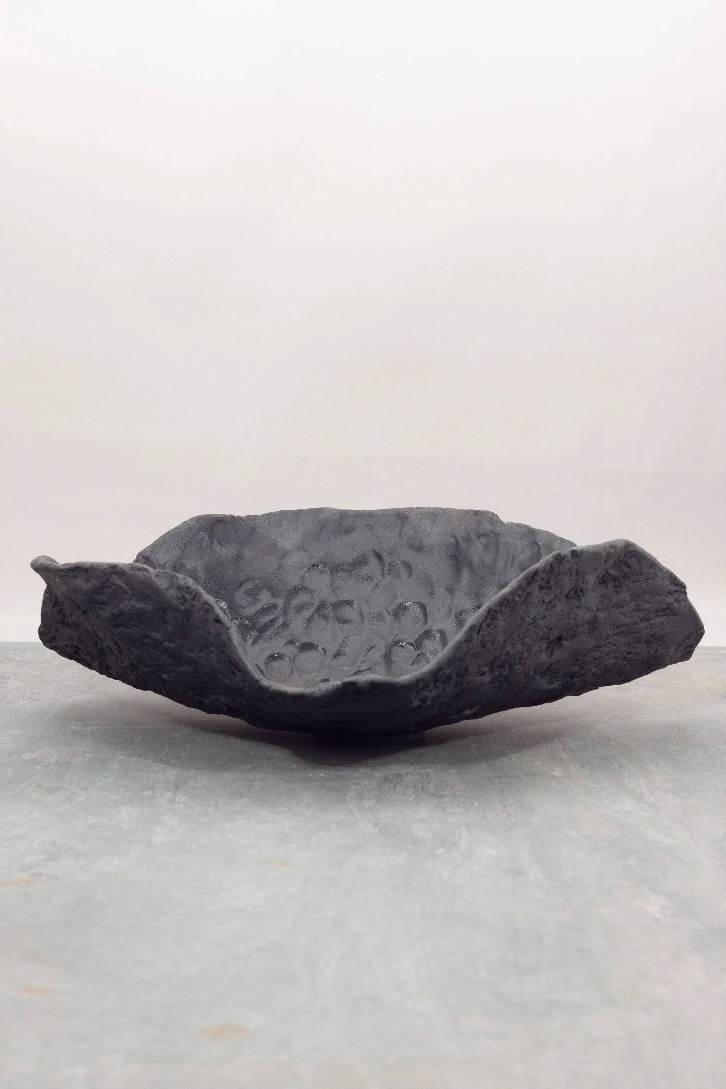 A black porcelain, textured, bowl-shaped sculpture with petal-like edges, placed on a flat surface against a plain, light-colored background.