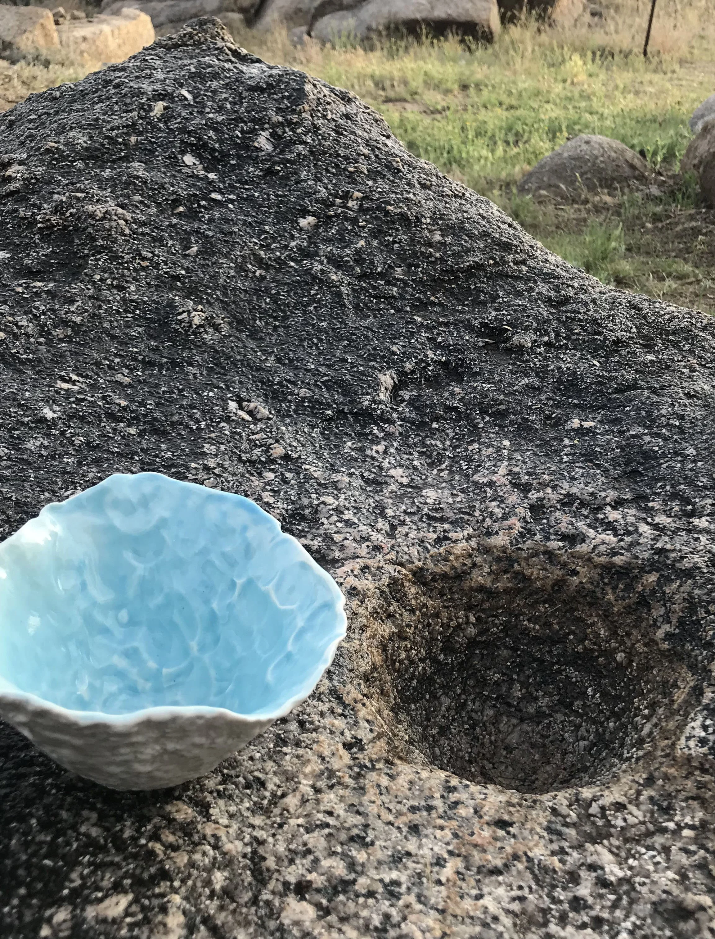 A close-up of a large granite rock that served as a grinding stone with a press molded porcelain bowl sitting on it, with grass in the background and additional rocks.