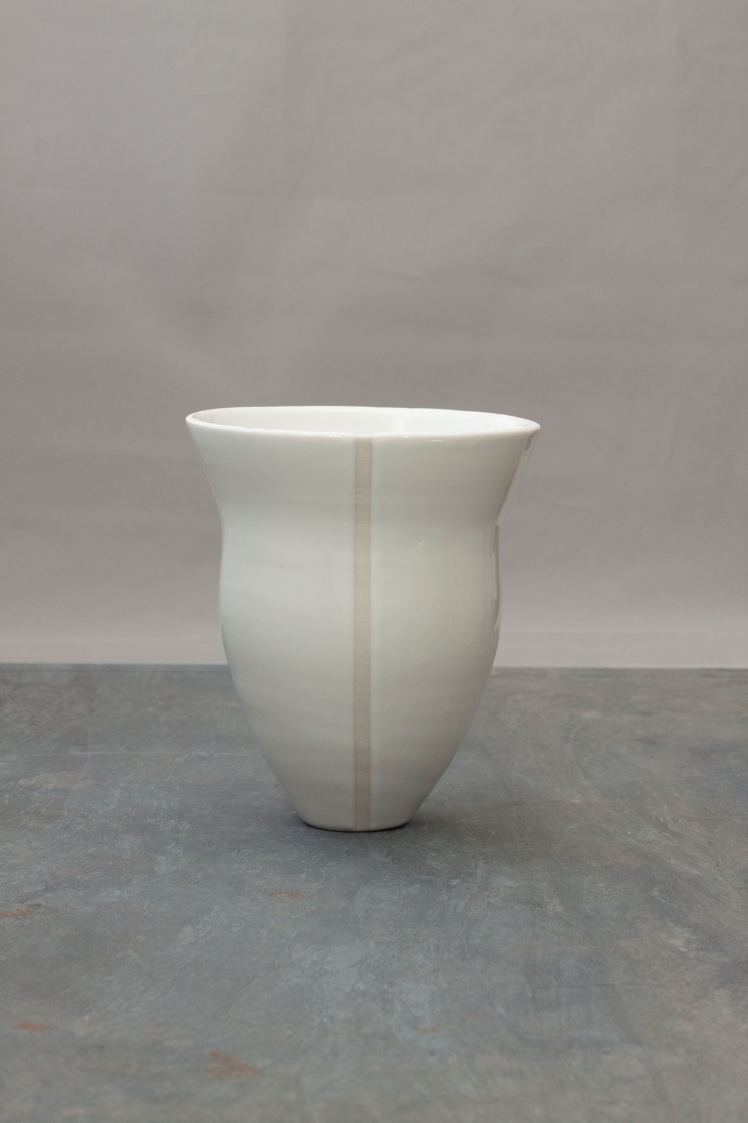 A minimalist white ceramic vase with a flared top and geometric shape, placed on a dark gray surface against a plain gray background.