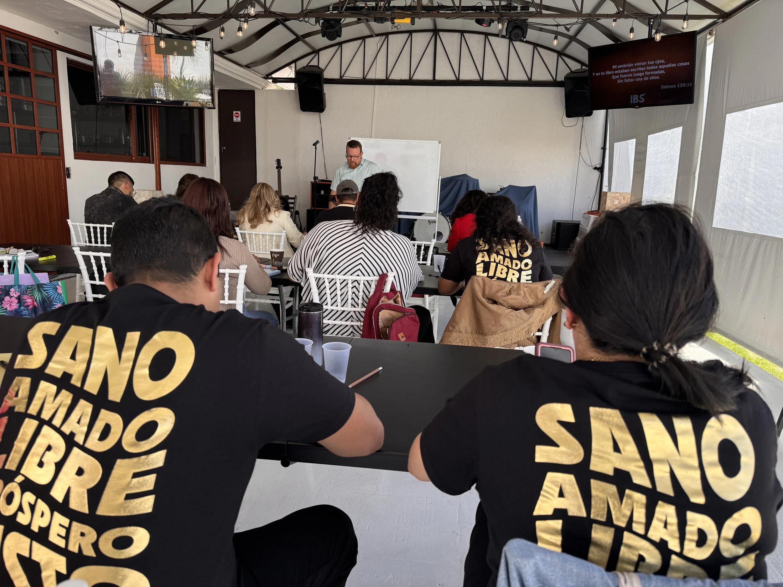 Grupo de personas en una clase o conferencia, algunas llevan camisetas negras con texto dorado. Un hombre está al frente junto a un pizarrón blanco. Hay pantallas y equipo de sonido en la sala.
