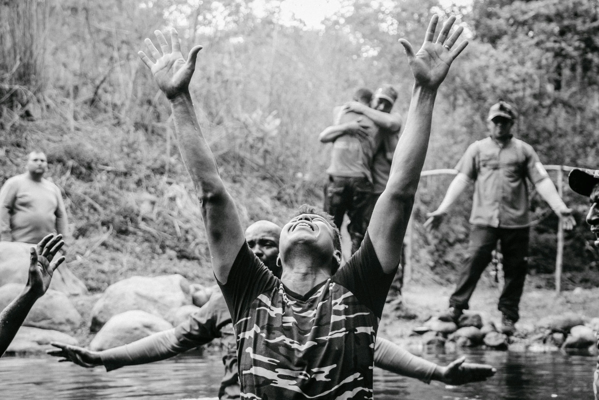 Un grupo de personas en una ceremonia al aire libre cerca de un río, una persona levantando los brazos y mostrando emoción, mientras otros observan y participan en el fondo.