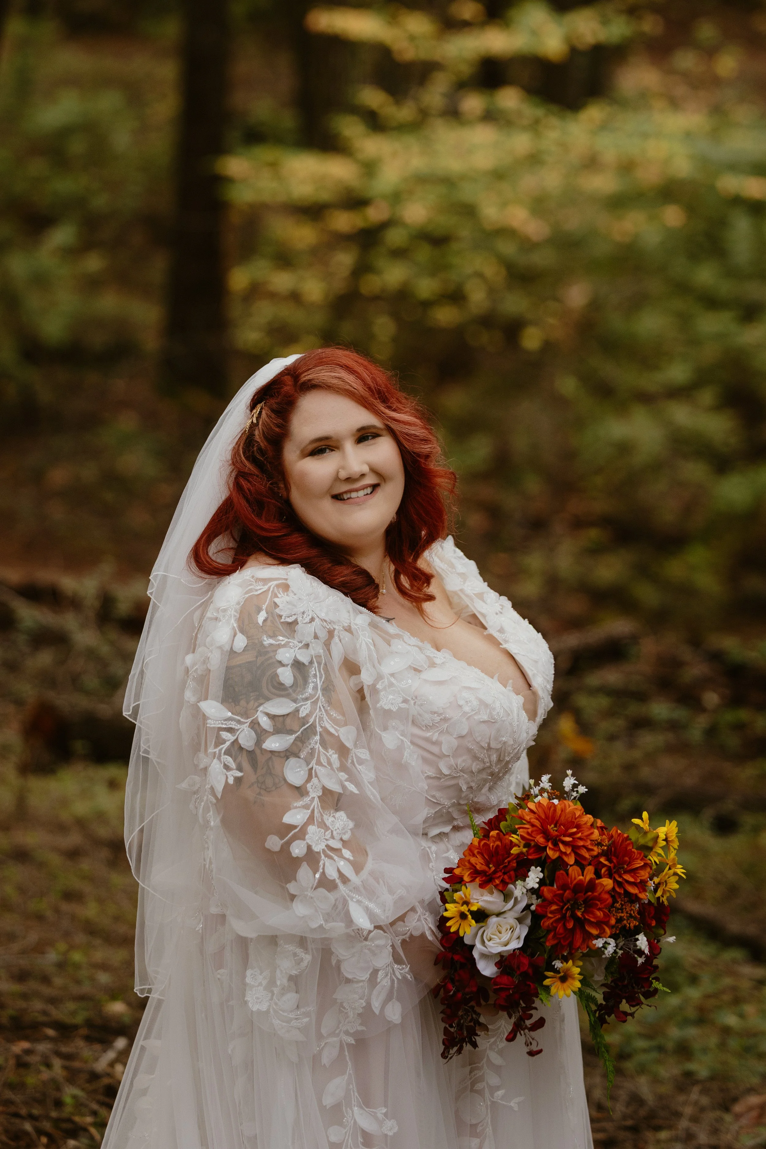 A woman with red hair wearing a lace wedding dress and veil, holding a colorful bouquet, standing outdoors in a forested area with autumn leaves.