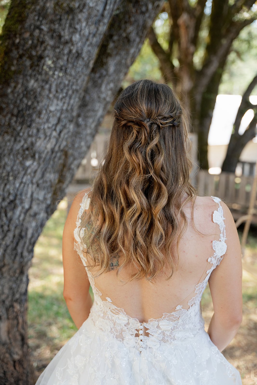 Back view of a woman with wavy, half-up hair standing outdoors in a white wedding dress with lace details.