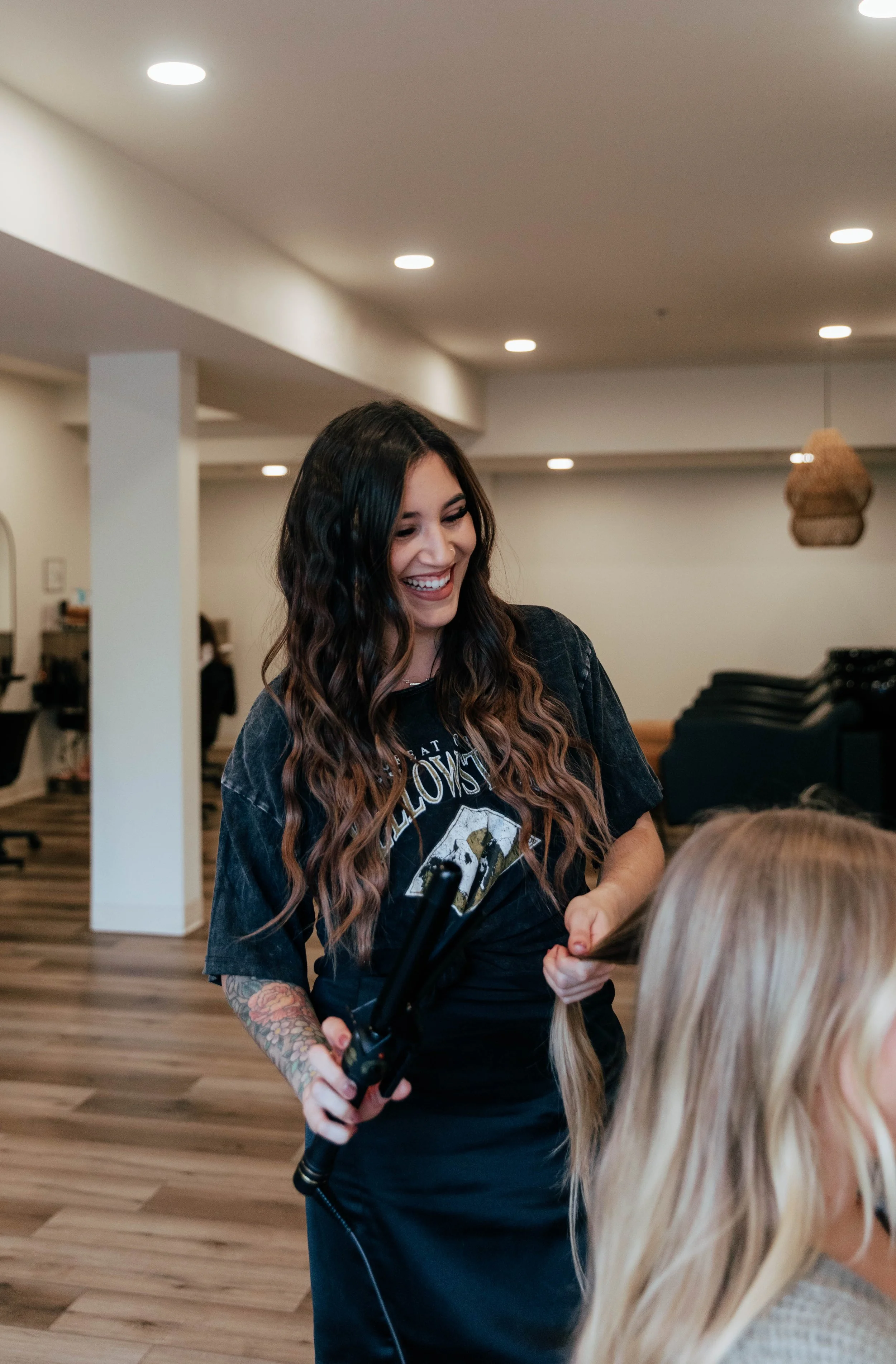 A smiling woman with long wavy brown hair and tattoos on her arm is holding a curling iron in a room with wooden flooring and hanging light fixtures. She is interacting with another woman with long blonde hair.