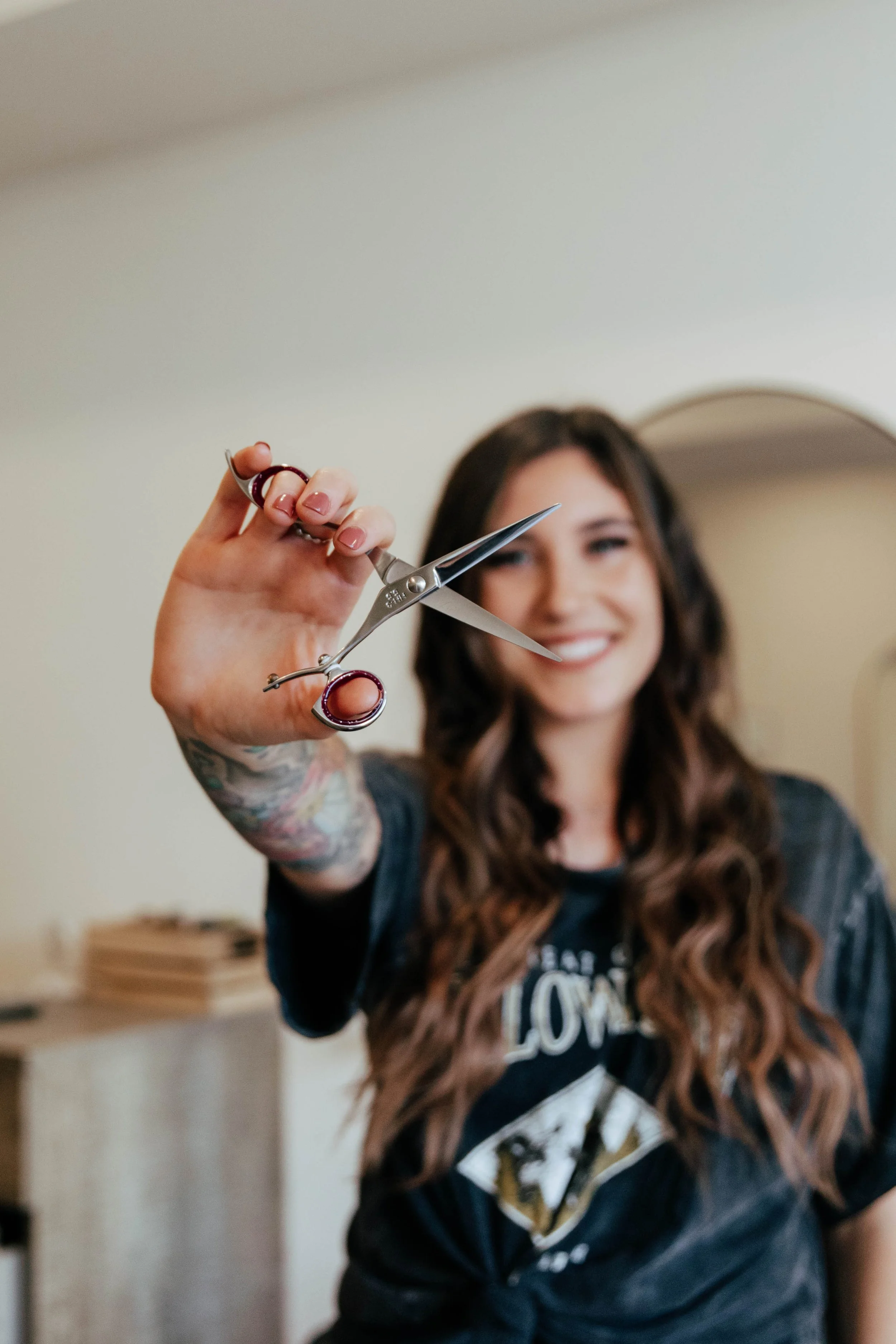 A woman with long, wavy brown hair and tattoos on her arm is smiling and holding a pair of scissors with red handles in front of her.