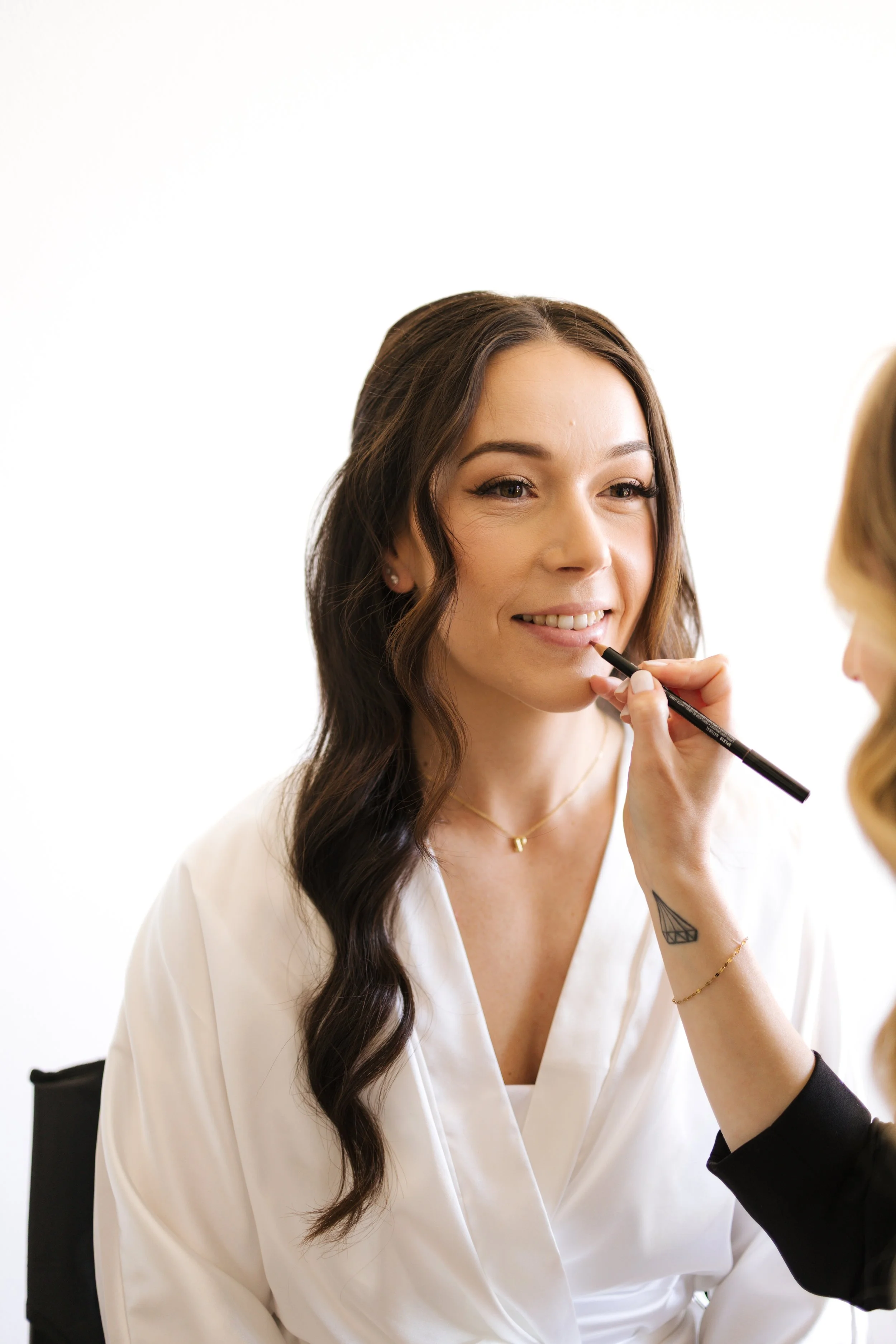 A woman with long wavy dark hair and light skin is getting her makeup done by a makeup artist. She is smiling, wearing a white shirt, and has a small tattoo on her wrist.