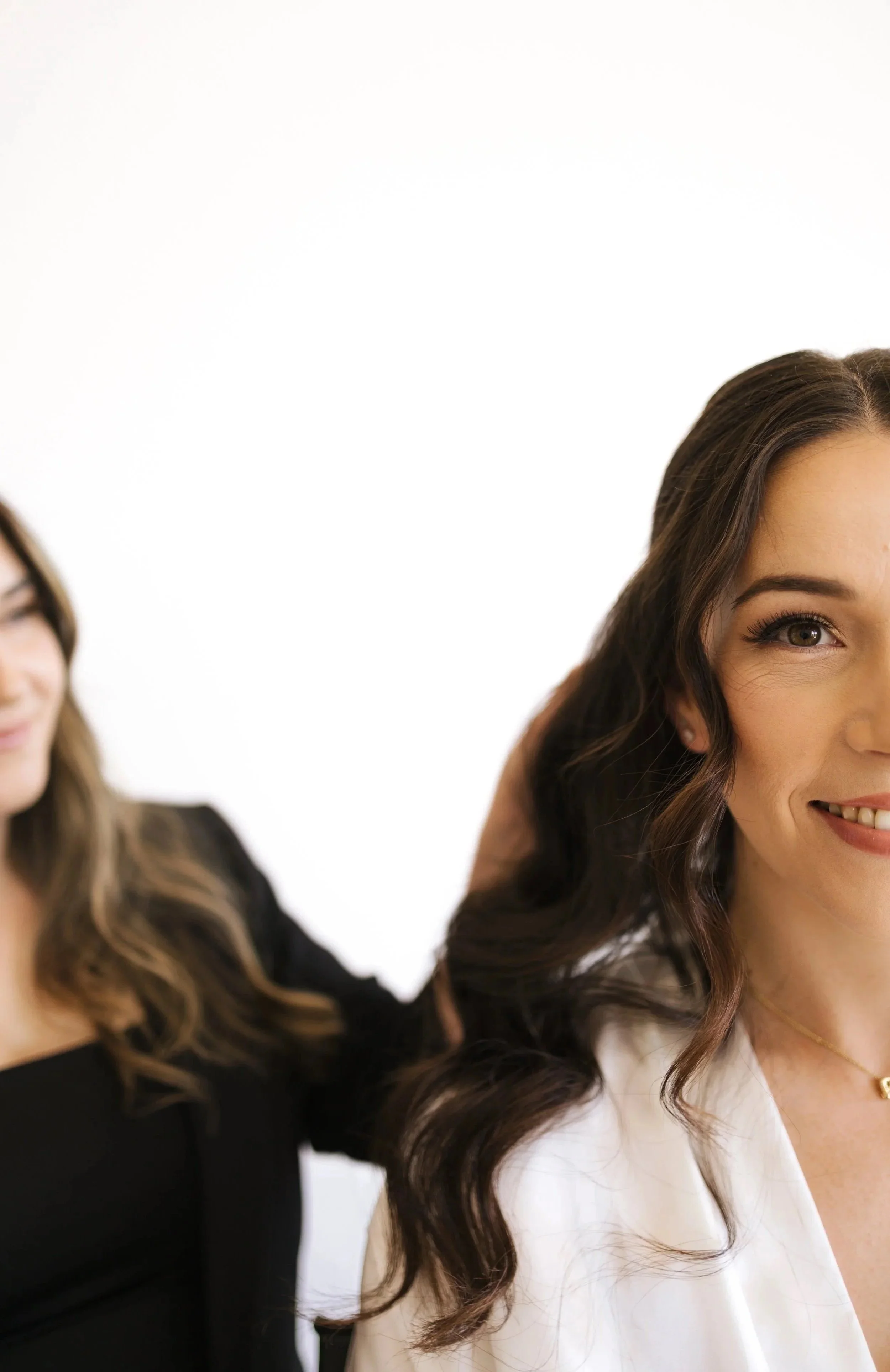Two women with long wavy hair, one with brown hair wearing a white top and makeup, the other with light brown hair and glasses wearing a black top, in front of a white background.