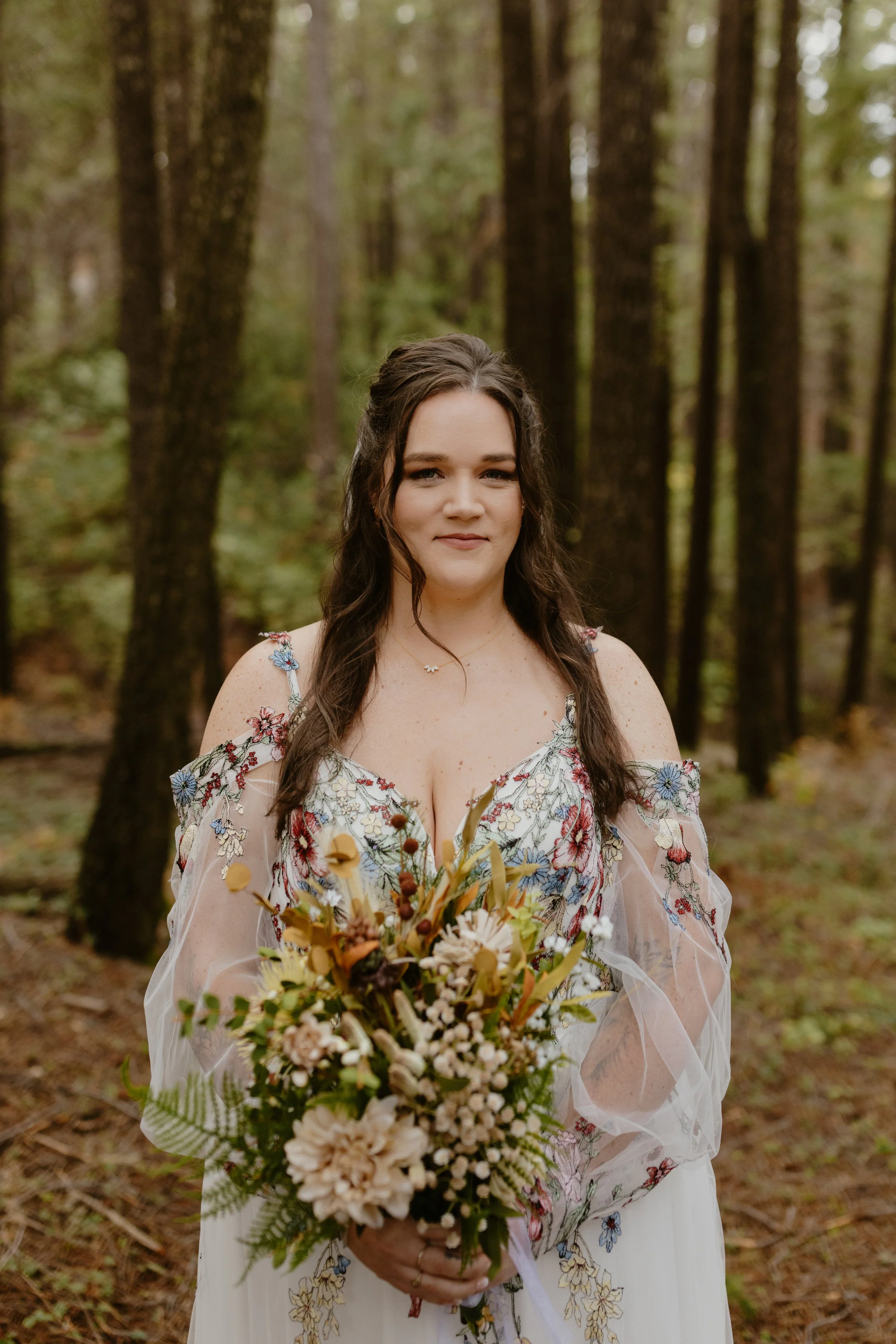 A woman in a floral dress holding a bouquet of flowers in a forest with tall trees and green foliage.