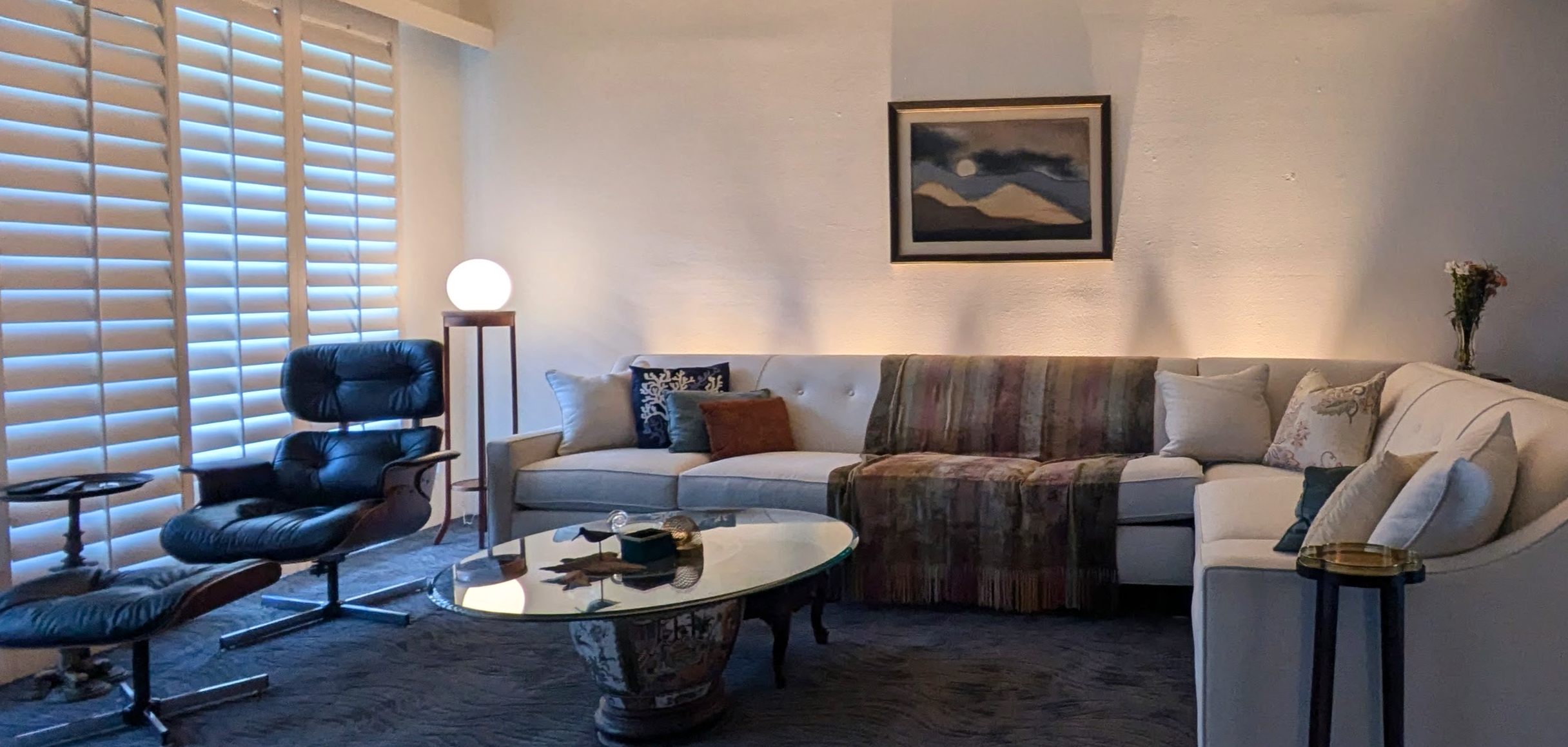 Living room with white sectional sofa, black lounge chairs, round glass coffee table, window with plantation shutters, framed landscape painting, floor lamp, and flower vase.
